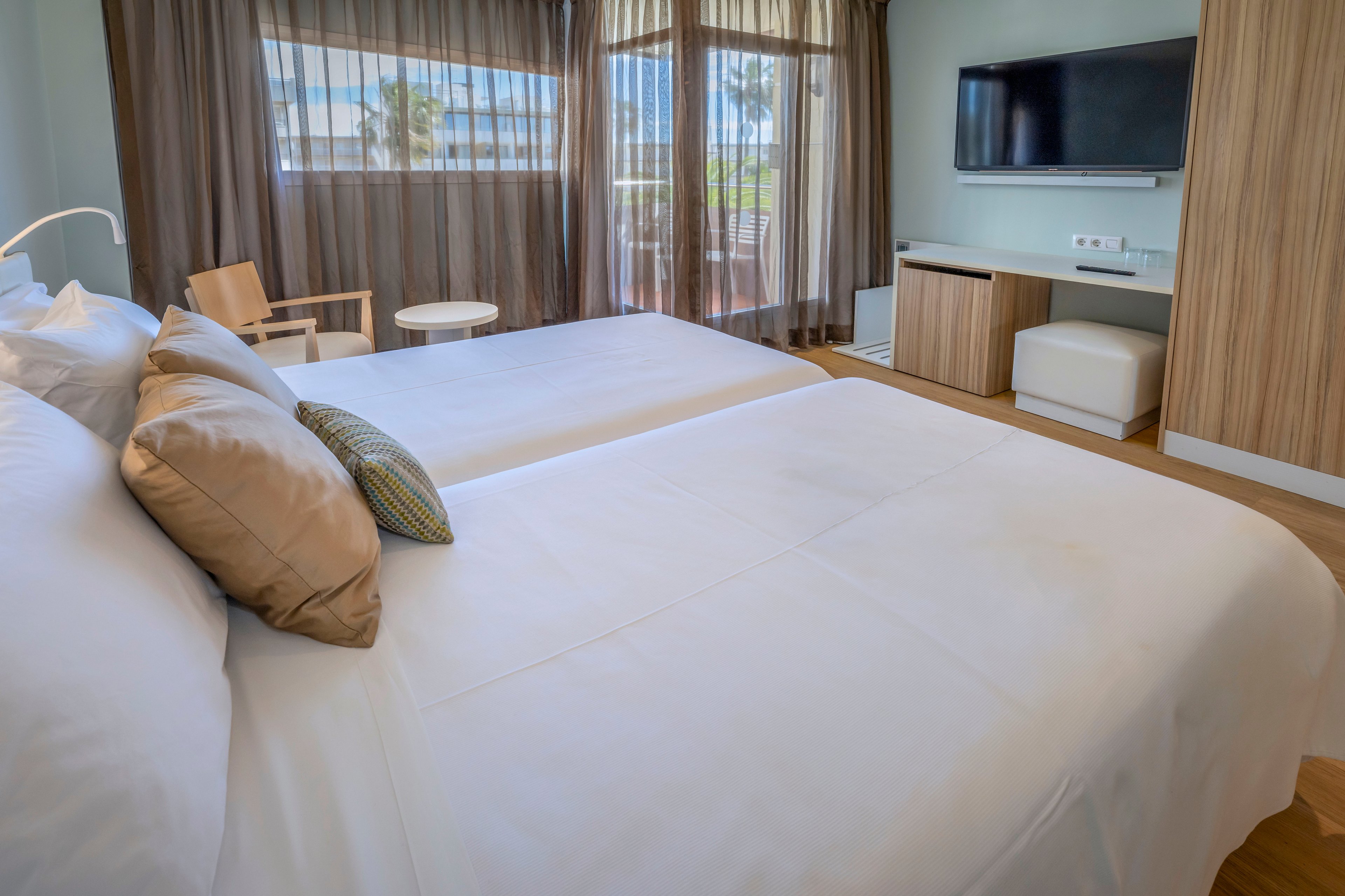 A contemporary hotel room features two neatly made twin beds with white linens and accent pillows. Floor-to-ceiling windows with sheer curtains allow natural light to brighten the space and reveal a partial outdoor view. A flat-screen television is mounted on the wall above a wooden cabinet and small ottoman. The room’s decor combines light wood furniture and soft neutral tones for a calm, minimalist atmosphere.