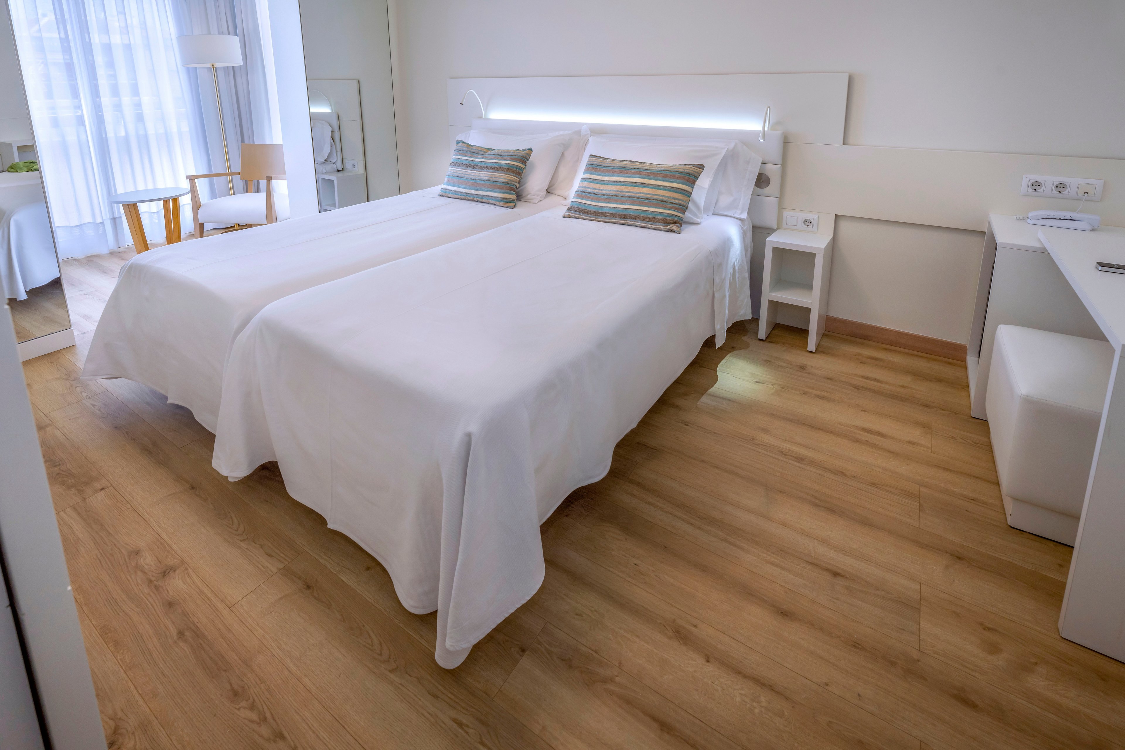 A contemporary hotel room features two neatly made twin beds with white linens and striped accent pillows. The space has light wood flooring, a white headboard with integrated lighting, and minimalist white furniture including a desk and bedside table. Natural light filters through sheer curtains covering a large window, illuminating a small seating area with a chair and side table. The overall design is clean, airy, and inviting, emphasizing comfort and simplicity.