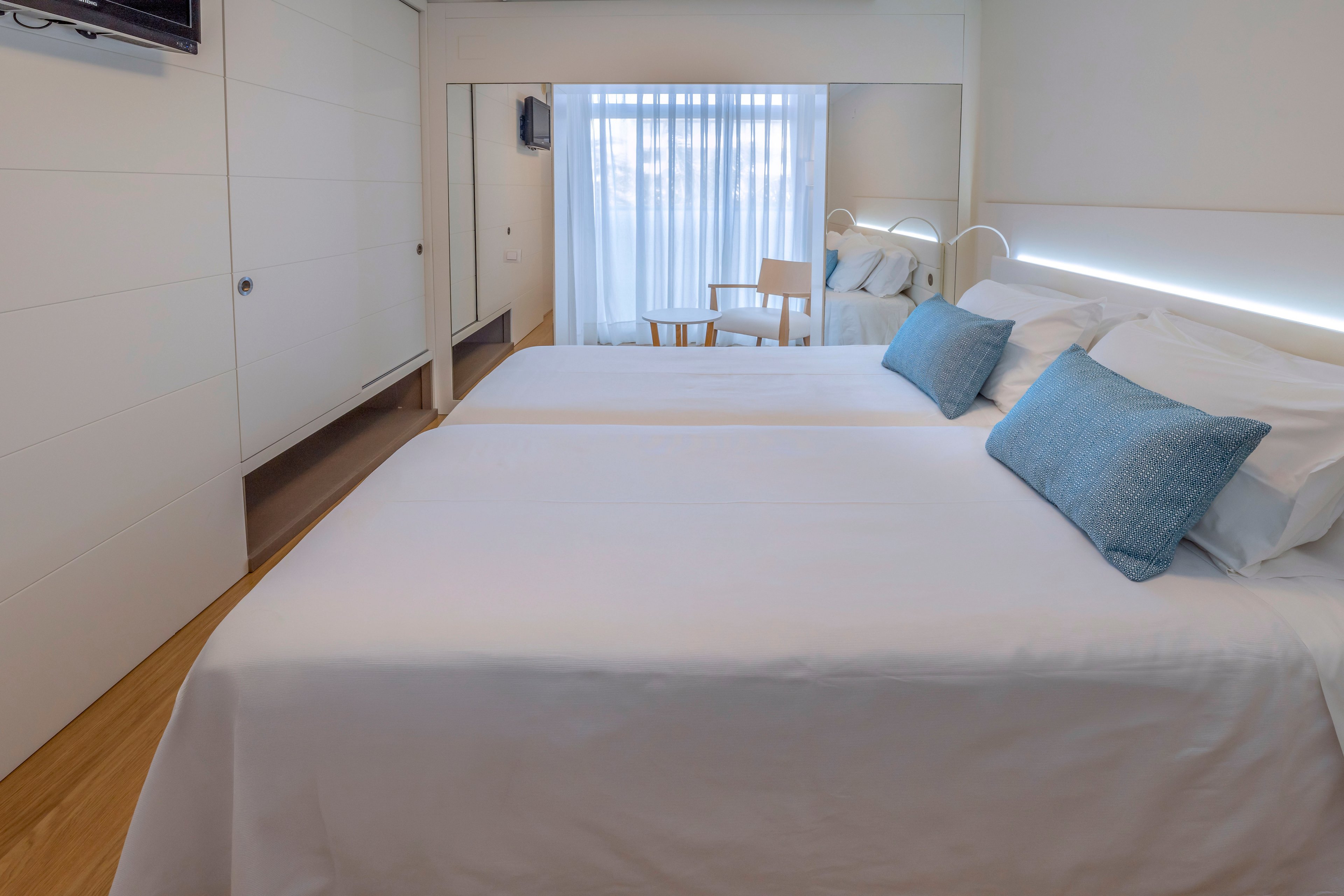A bright modern bedroom features two neatly made twin beds with crisp white linens and light blue accent pillows. The room has a minimalist design with white built-in wardrobes and a large mirror reflecting the space. Natural light filters through sheer curtains covering a floor-to-ceiling window or balcony door. A simple wooden chair and small table sit near the window, enhancing the clean and airy atmosphere.