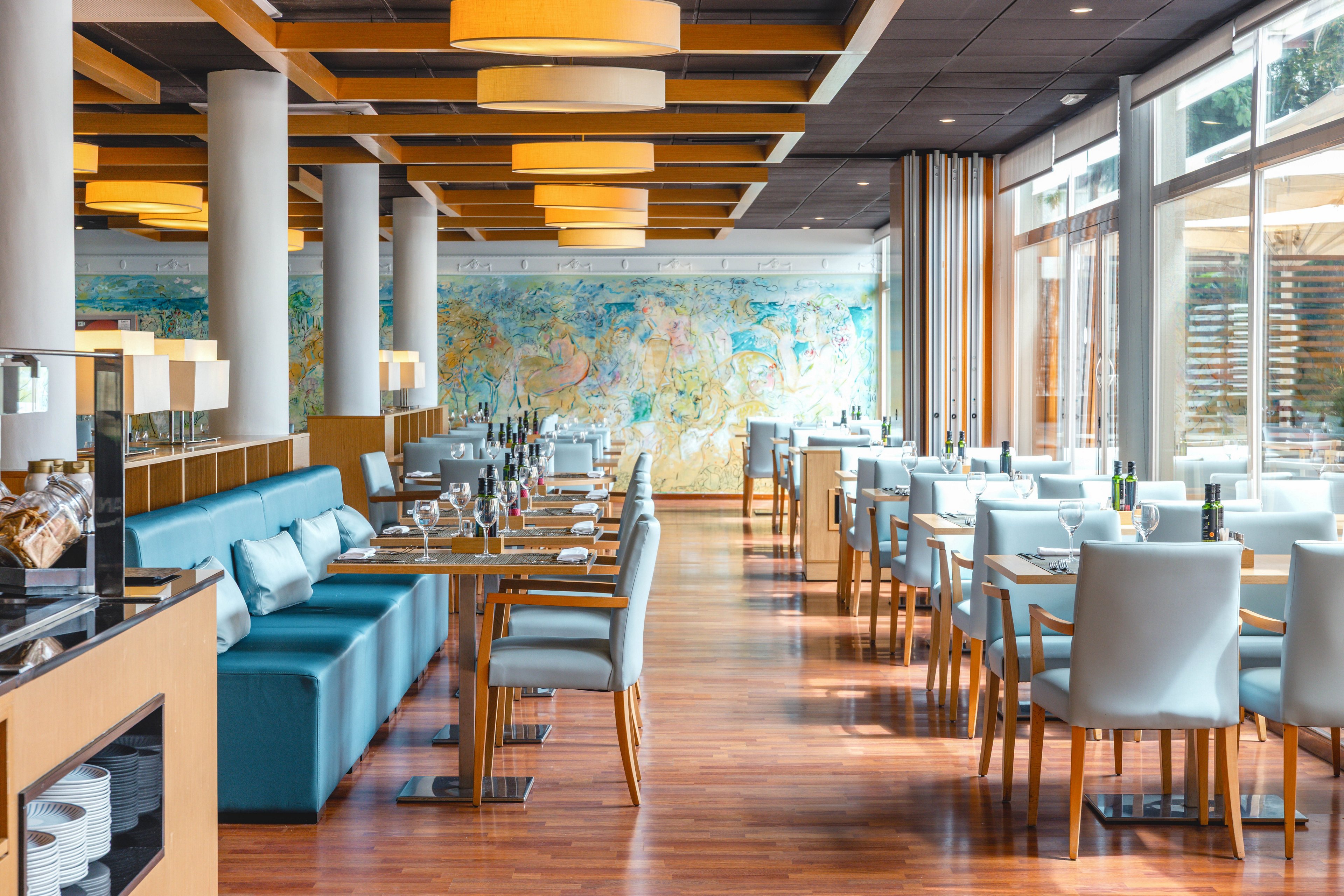 A contemporary restaurant dining room features neatly arranged tables with light blue chairs and banquette seating. The space is bright and airy, with large windows along one side letting in natural light. Wooden floors and ceiling beams add warmth, while cylindrical pendant lights create a modern ambiance. Tables are set with glassware, cutlery, and bottles, ready to welcome guests. A colorful mural spans the back wall, adding artistic interest to the interior.
