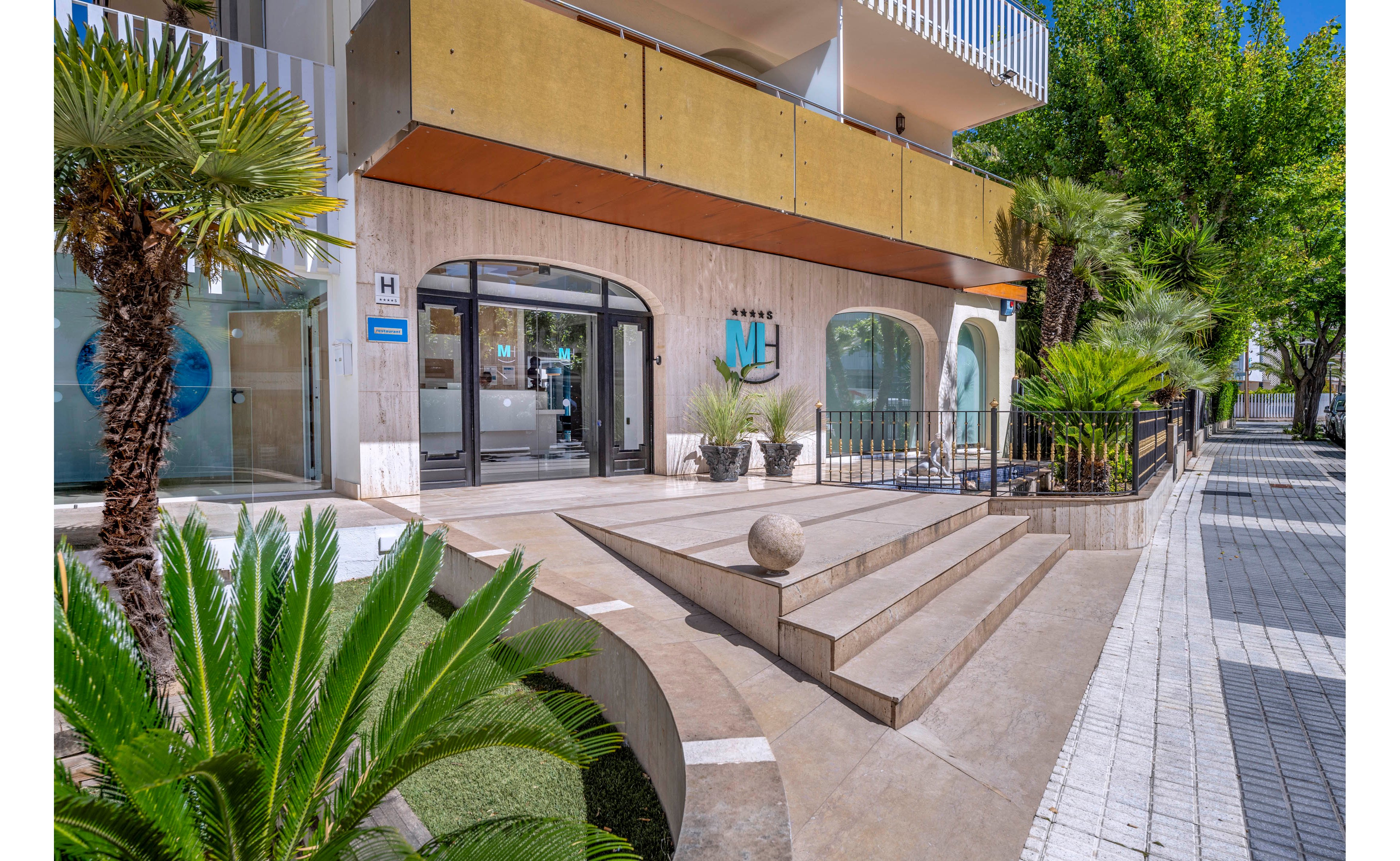 A contemporary hotel entrance features wide steps leading up to glass doors and a minimalist facade. Palm trees and other greenery frame the walkway, adding a tropical touch to the urban sidewalk. Large windows reveal the bright interior, while balconies and geometric railings accent the exterior architecture. The scene is captured on a sunny day with clear blue skies and strong natural light.