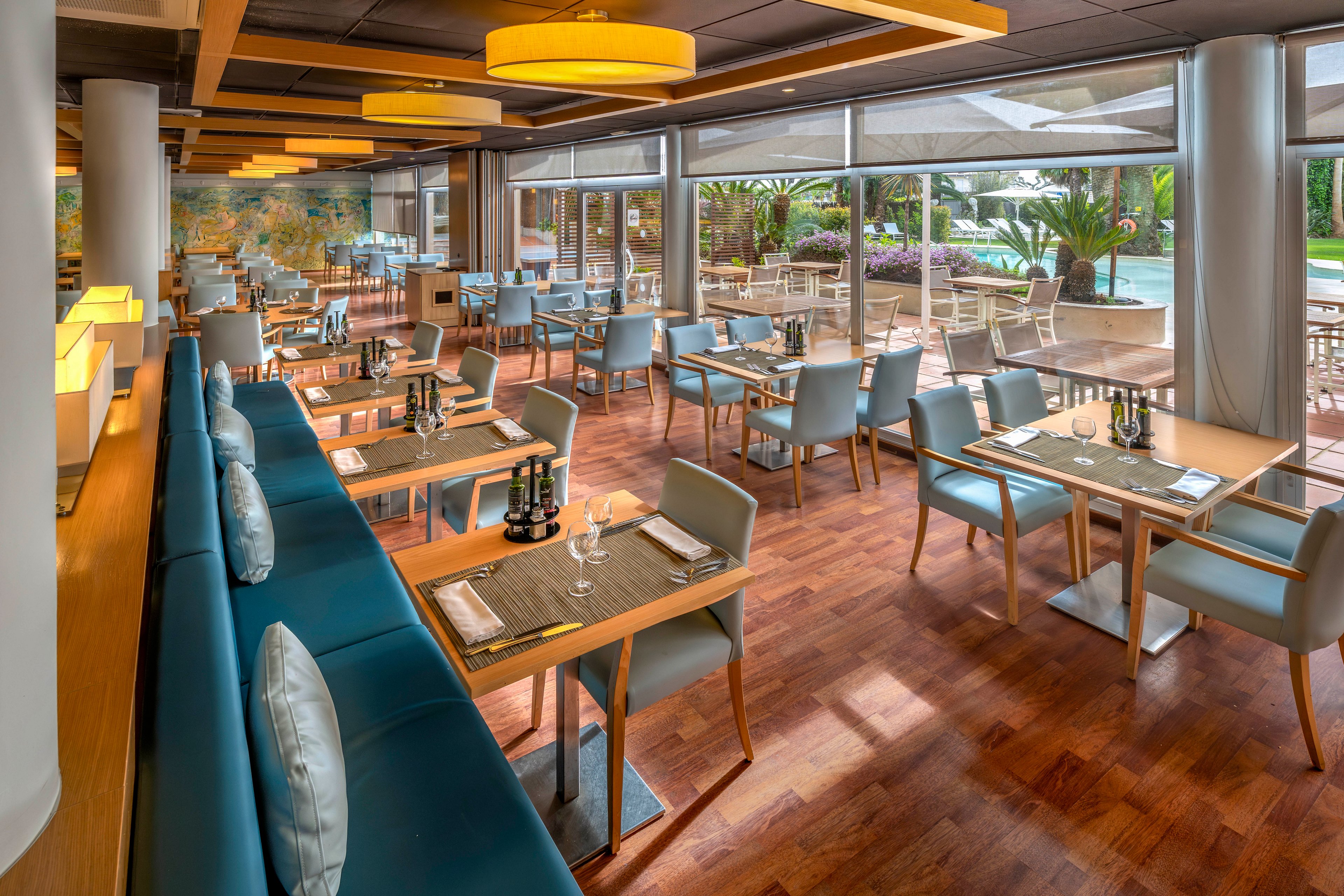 A contemporary restaurant interior features neatly arranged tables and cushioned bench seating. Large windows open onto an outdoor terrace with additional dining tables and views of a landscaped pool area. Warm wooden flooring and ceiling accents contrast with cool blue chairs and neutral tones. Soft pendant lighting and natural daylight create an inviting, relaxed dining atmosphere.