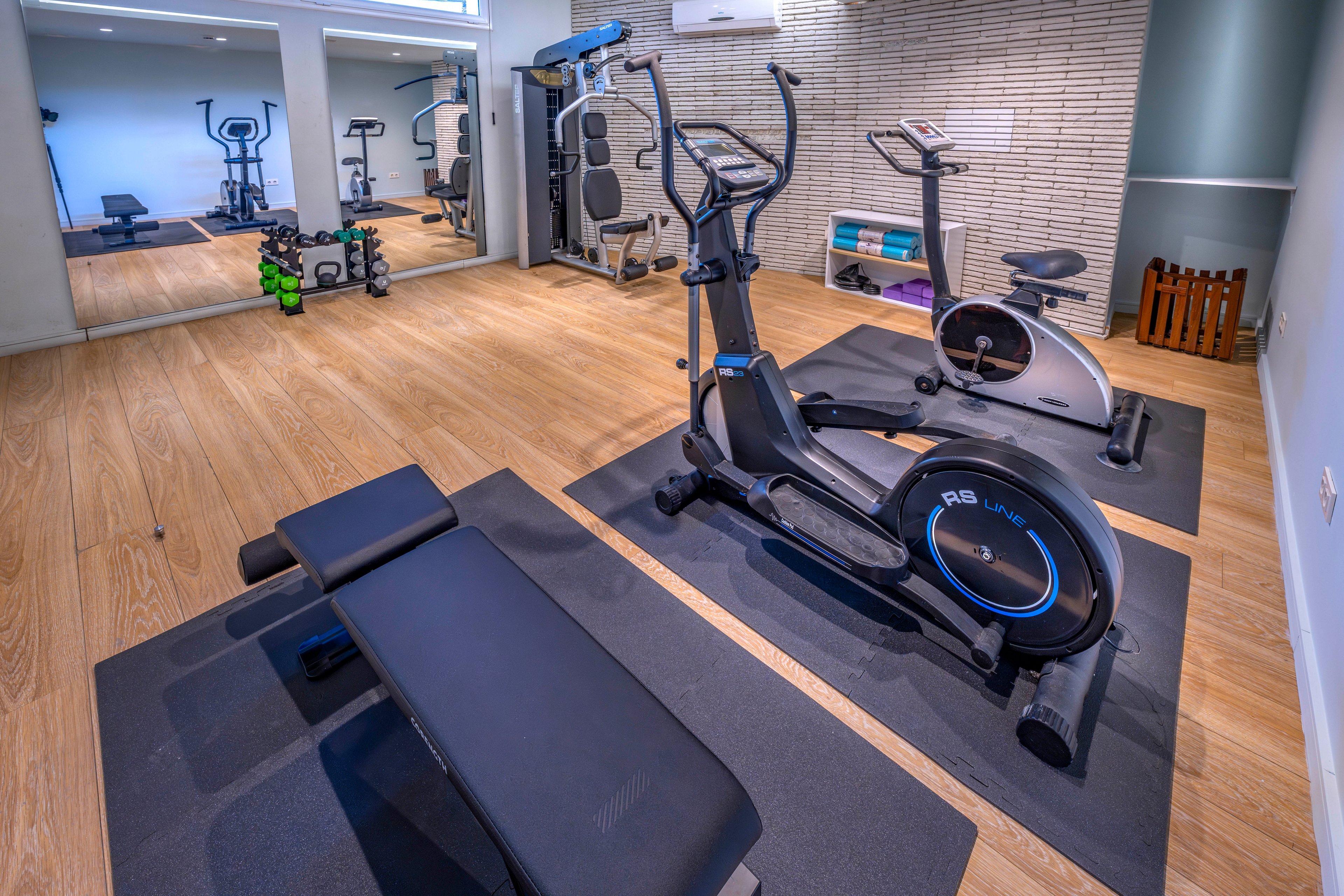 A modern home gym features a stationary bike, elliptical trainer, and weight bench arranged on black mats. The room has light wooden flooring and a neutral-toned wall with a large mirror reflecting the equipment. Additional free weights and a multi-gym machine are visible in the background, creating a complete workout space. Bright, even lighting and clean lines give the room a contemporary, minimalist feel.