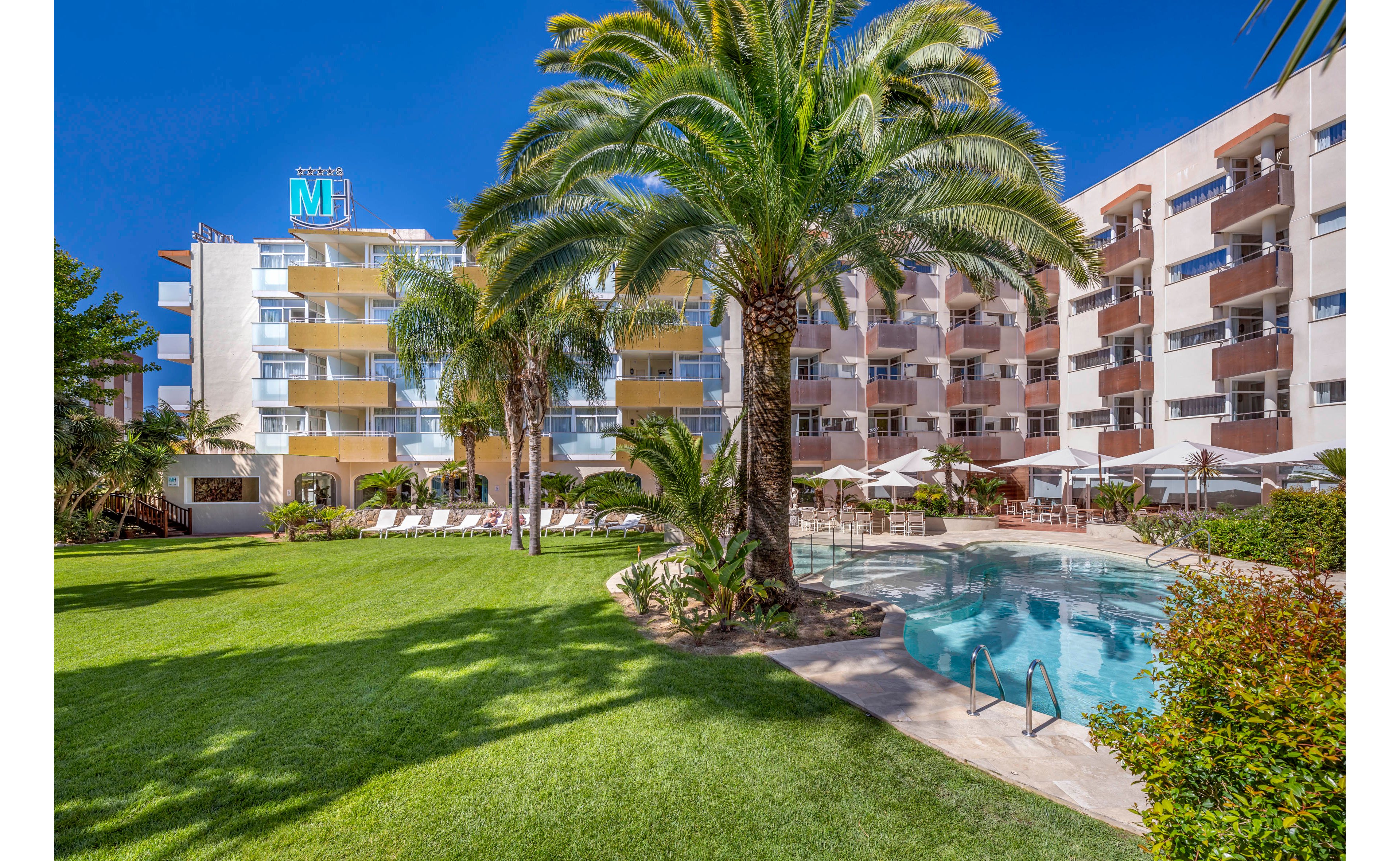 A contemporary multi-story hotel overlooks a curved outdoor swimming pool surrounded by sun loungers and umbrellas. Lush green lawns and tall palm trees create a tropical garden atmosphere in the courtyard. The building features balconies on each floor, offering views of the pool and landscaped grounds. Clear blue skies and bright sunlight enhance the relaxed vacation setting.