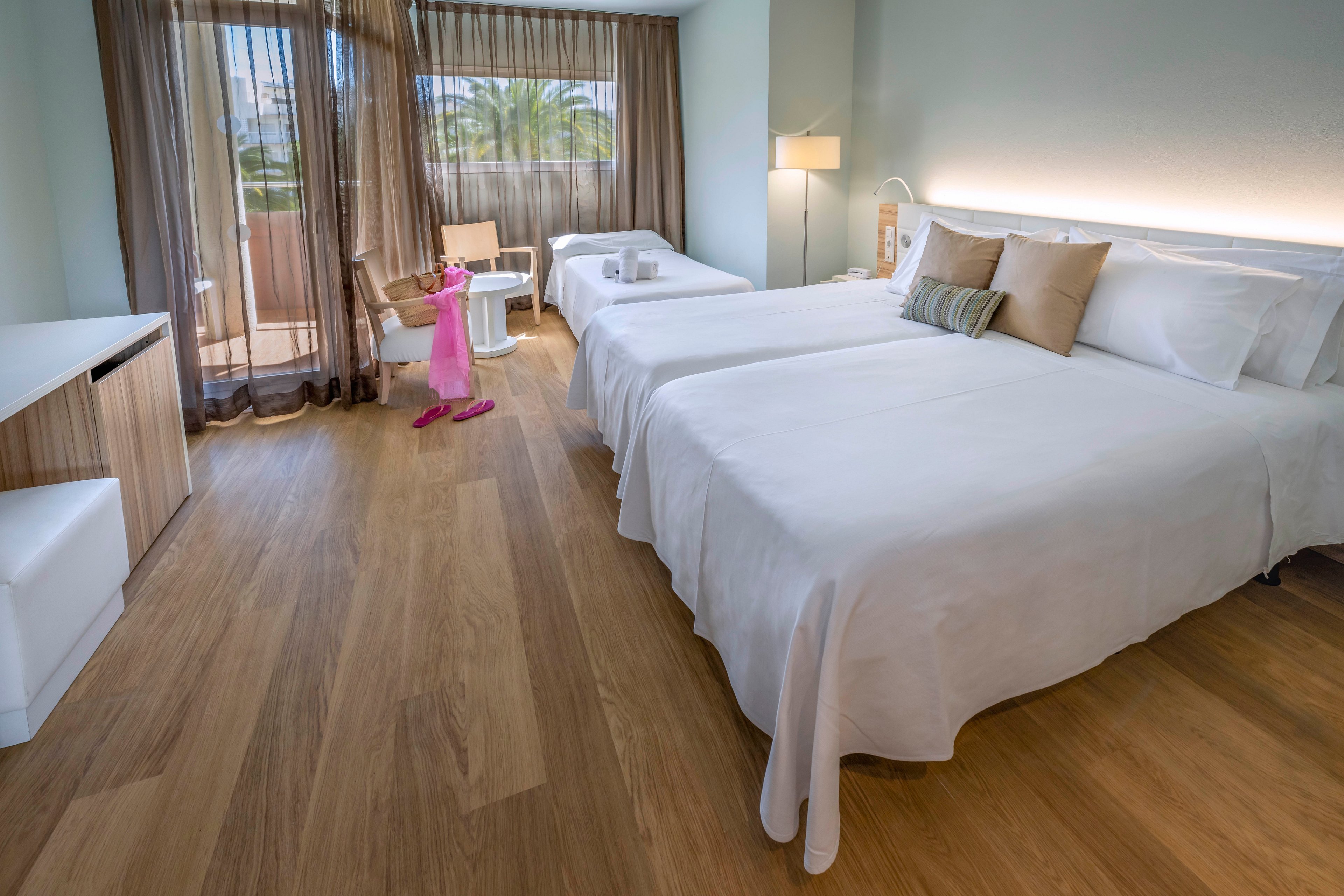 A bright, modern hotel room features two neatly made beds with white linens and neutral accent pillows. The space has warm wooden flooring, a desk area, and a small table with a pink dress draped over a chair. Floor-to-ceiling windows with sheer curtains open to a balcony, revealing palm trees outside. Soft lighting and minimalist decor create a calm, inviting atmosphere suitable for vacation or business travel.