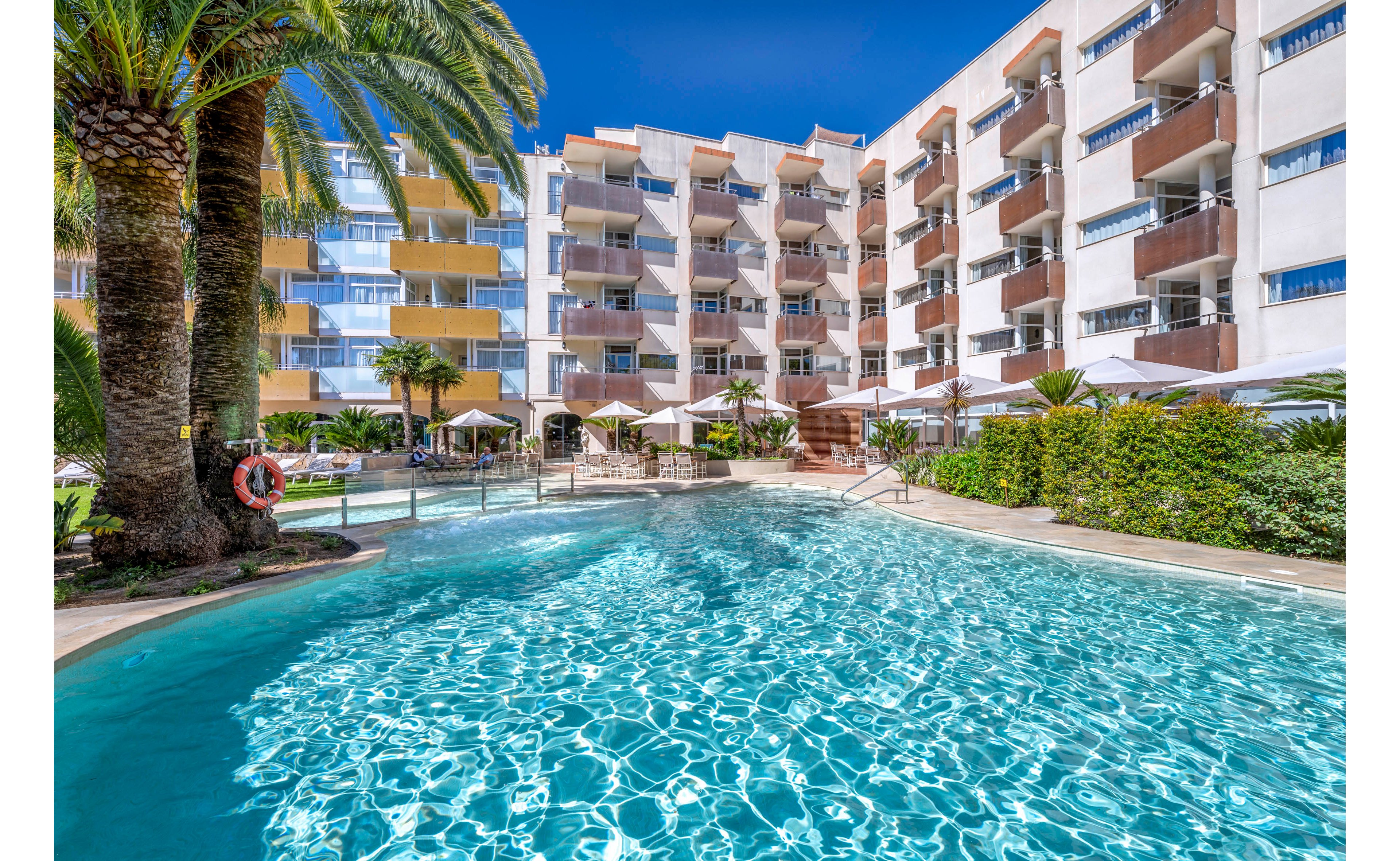 A large outdoor swimming pool sparkles under a clear blue sky in front of a modern multi-story resort hotel. Palm trees and lush greenery frame the pool area, creating a tropical vacation atmosphere. Sun loungers and umbrellas line the poolside terrace, suggesting a relaxing holiday setting. The building’s balconies overlook the water, offering guests views of the pool and landscaped courtyard.