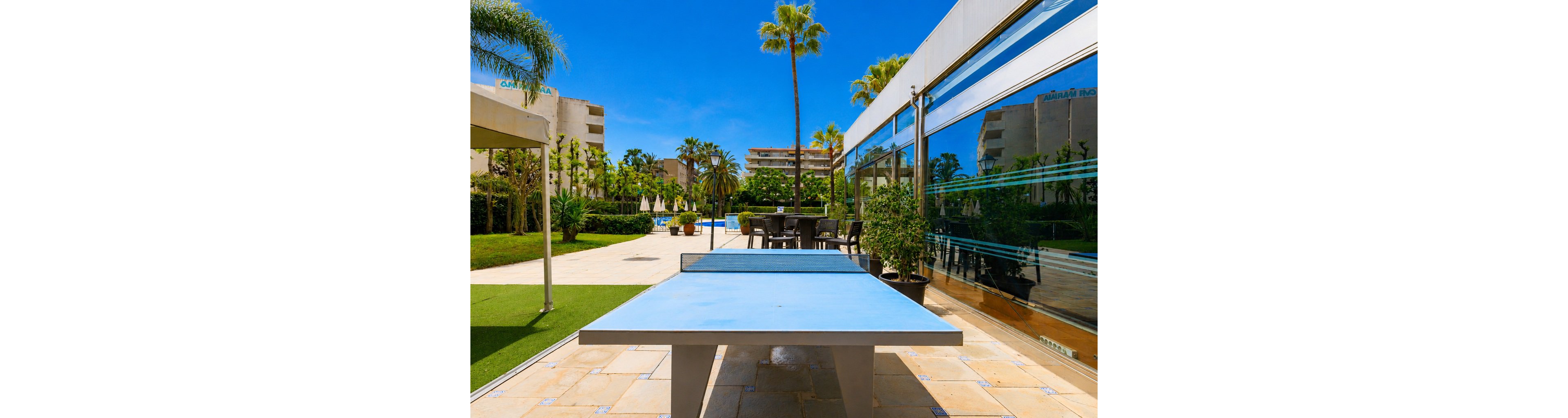An outdoor ping pong table stands in the foreground of a sunny resort courtyard. Behind it, a swimming pool area with umbrellas, lounge chairs, and palm trees creates a vacation atmosphere. A modern glass-front building reflects the bright blue sky and surrounding greenery. The scene suggests leisure, recreation, and relaxation in a warm climate.