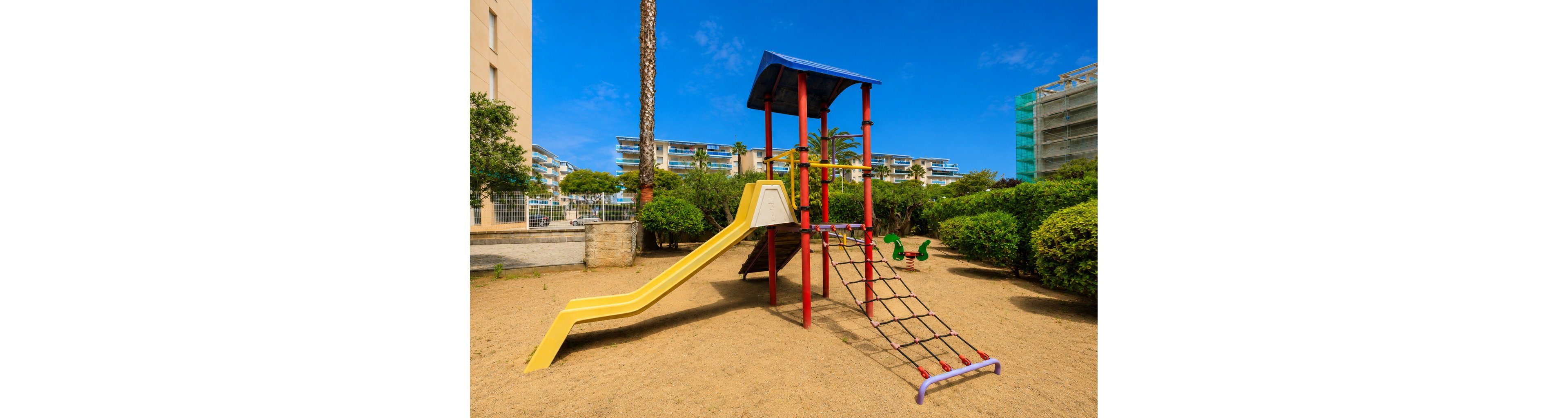 A colorful playground structure with a yellow slide and rope climbing net stands on a sandy surface in an outdoor courtyard. The playset features red and blue support beams and a shaded roof, with additional spring riders visible in the background. Green bushes, palm trees, and modern apartment buildings surround the play area under a bright blue sky. The scene suggests a sunny, family-friendly environment designed for children’s recreation.
