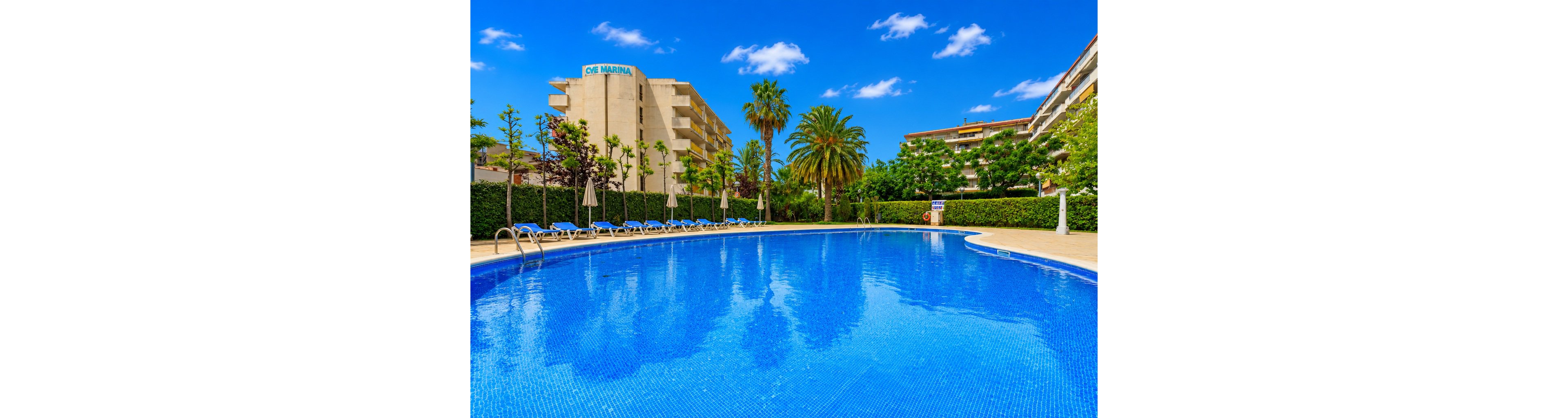A large curved outdoor swimming pool glistens under a bright blue sky with scattered clouds. Sun loungers line the poolside, facing the water and bordered by manicured hedges and trees. Tall palm trees and lush greenery frame the scene, with multi-story resort buildings visible in the background. The atmosphere is calm and inviting, suggesting a warm, relaxing vacation setting.