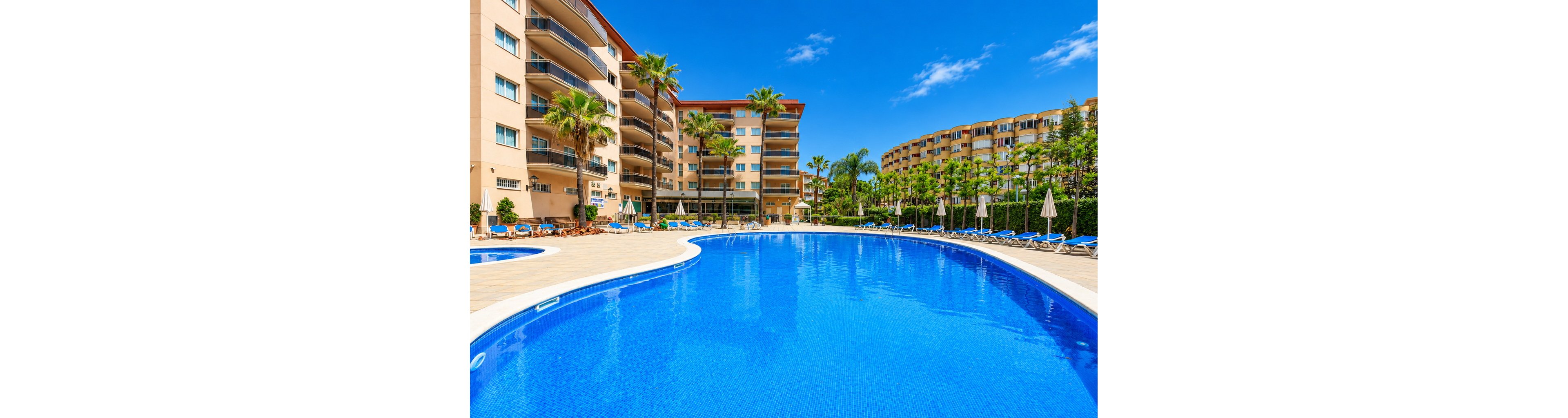 Large curved outdoor swimming pool is surrounded by sun loungers and parasols in a bright resort setting. Multi-story apartment buildings with balconies frame the pool area, with palm trees and lush greenery adding a tropical feel. The clear blue water reflects the vivid sky, creating a vibrant vacation atmosphere. The scene suggests a quiet, sunny day at a holiday complex.