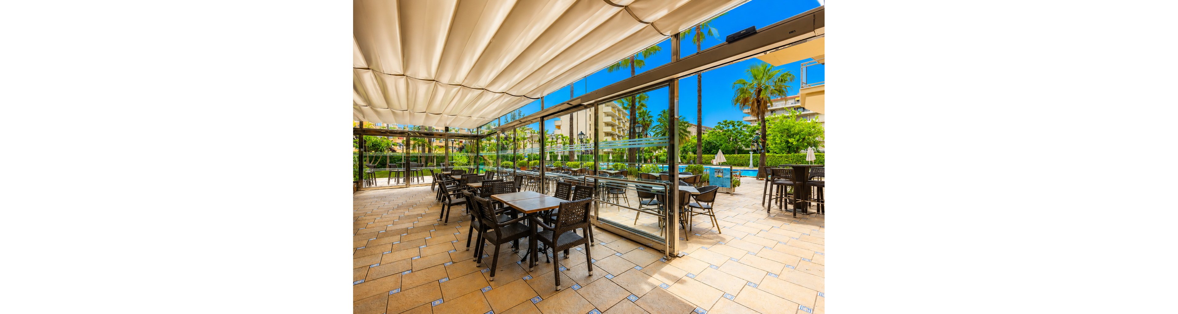 Spacious outdoor dining area with dark wicker chairs and tables sits beneath a large retractable canopy. Glass walls open to a lush garden with palm trees and a swimming pool in the background. The bright blue sky and warm sunlight create a relaxed, vacation-like atmosphere. The tiled floor and modern furnishings suggest a contemporary hotel or resort setting.