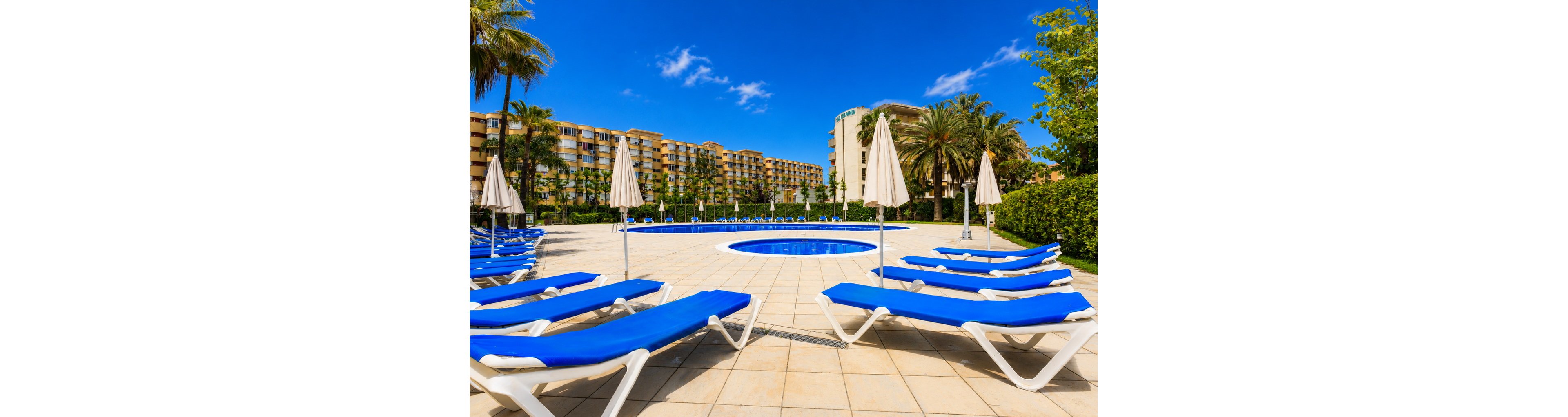 An outdoor resort pool area features rows of blue sun loungers arranged around a circular pool. Closed white umbrellas stand between the loungers on a wide tiled terrace. In the background, a large multi-story hotel building and lush palm trees frame the scene. The bright blue sky and vivid colors create a warm, inviting vacation atmosphere.