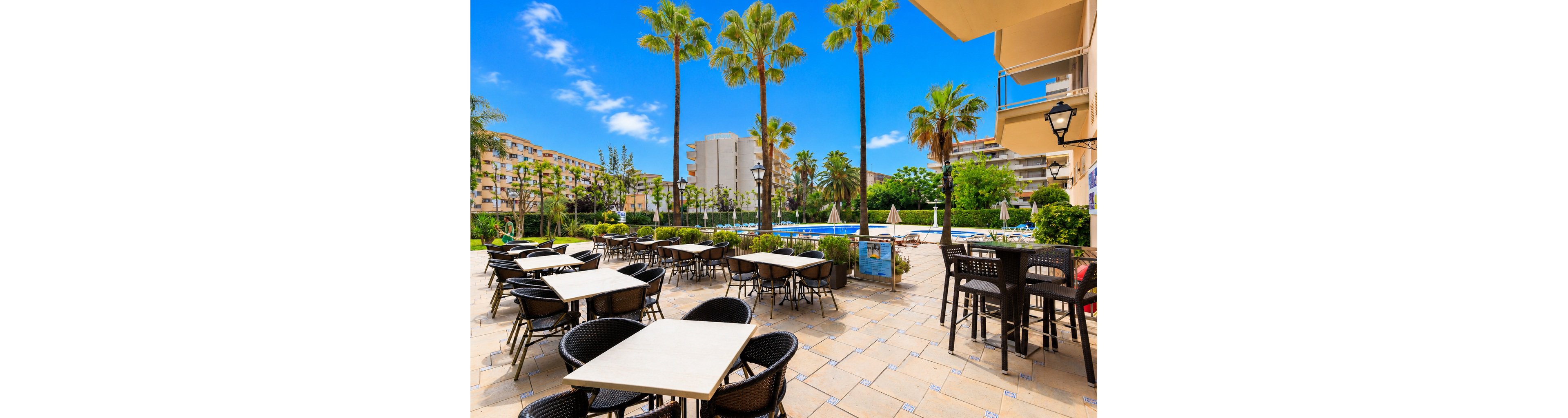An empty hotel terrace with tables and wicker chairs faces a large outdoor swimming pool. Tall palm trees line the pool area, with sun loungers and umbrellas arranged around the water. Apartment-style buildings surround the lush green garden, creating a resort-like atmosphere. The bright blue sky and vivid colors suggest a warm, sunny day ideal for relaxing outdoors.