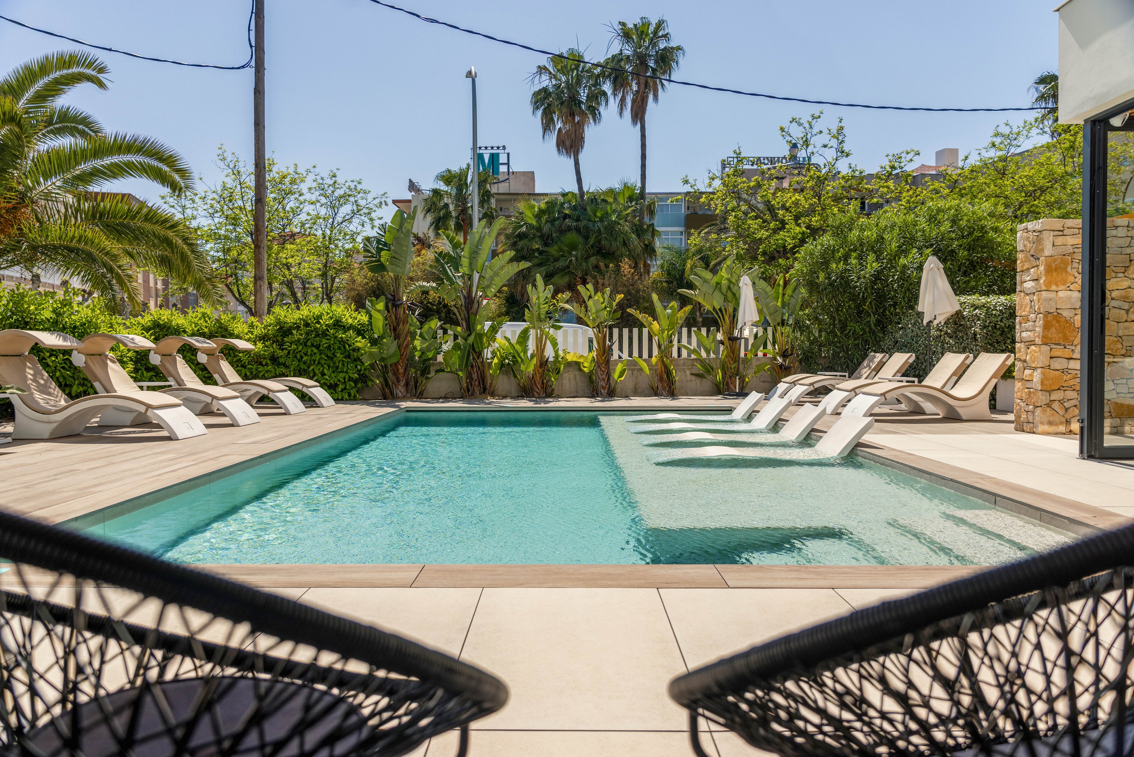 A rectangular outdoor swimming pool is surrounded by numerous reclining lounge chairs under a clear blue sky. The scene appears to be part of a hotel or resort courtyard, framed by lush greenery and palm trees. Two modern woven chairs in the foreground look toward the water, creating a relaxed vacation atmosphere. Buildings and umbrellas in the background suggest a comfortable, sunny retreat for guests.