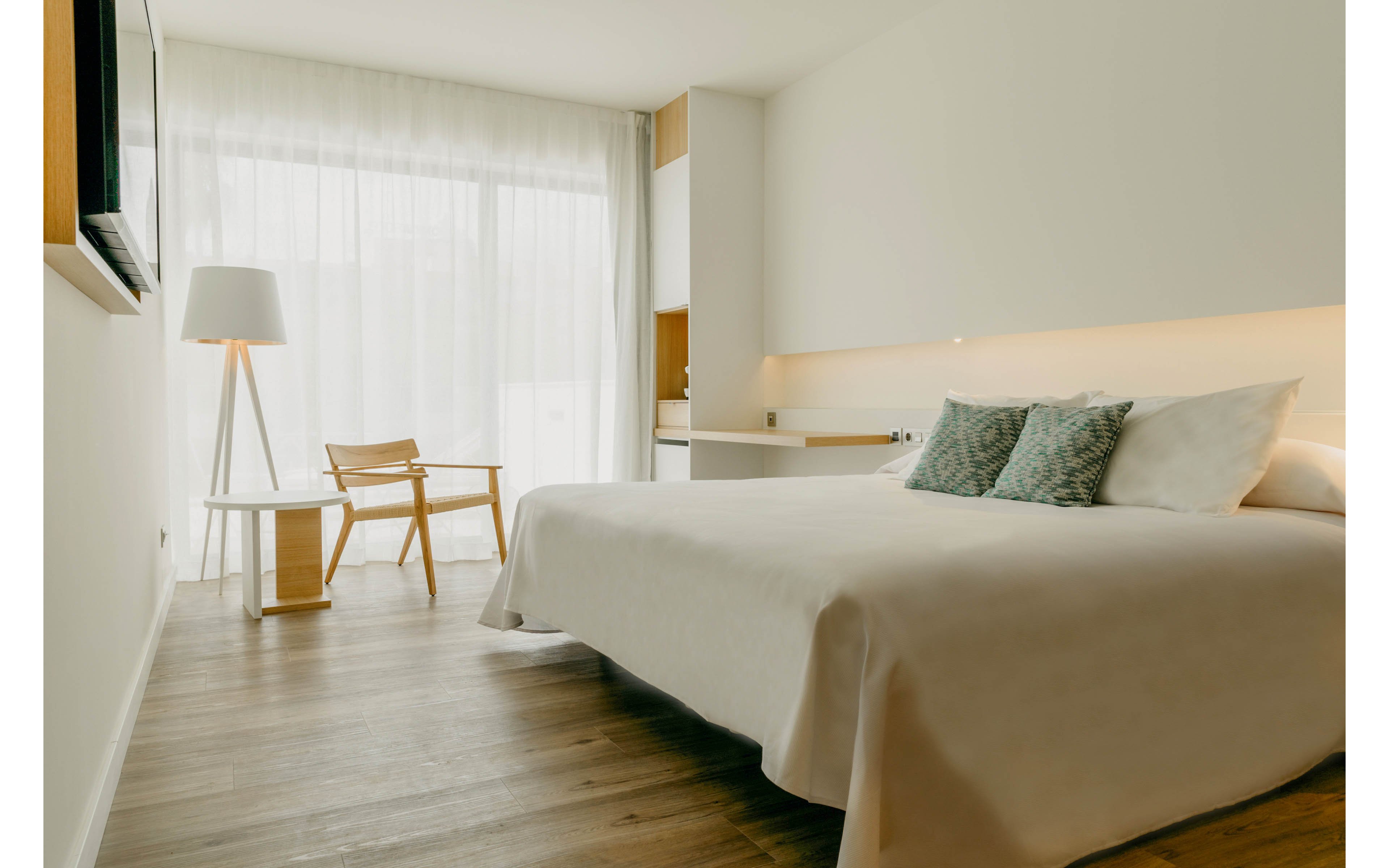 A bright, minimalist bedroom features a large neatly made bed with light-colored linens and patterned pillows. Floor-to-ceiling sheer curtains allow soft natural light to fill the space. A simple wooden chair and round side table sit near a tall floor lamp, enhancing the clean, modern aesthetic. Warm wooden flooring contrasts gently with the white walls and furniture, creating a calm and inviting atmosphere.