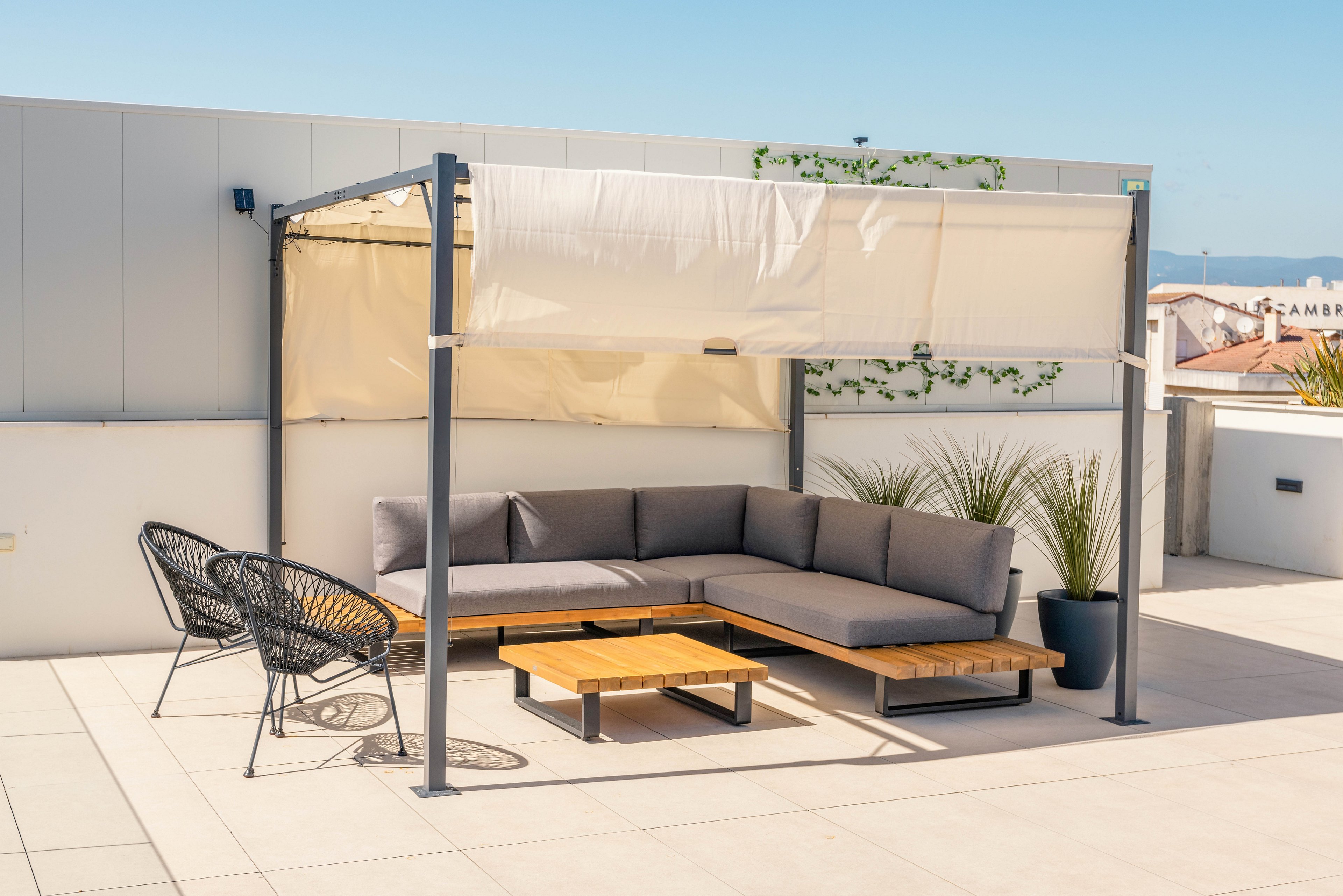 A contemporary rooftop patio features a shaded lounge area with a sectional outdoor sofa and low wooden tables beneath a minimalist pergola. Two black woven chairs sit nearby on the light tiled floor, creating a relaxed seating arrangement. Potted plants and a small green vine on the wall add a touch of nature to the clean, urban setting. Bright daylight and clear skies emphasize the airy, modern atmosphere.