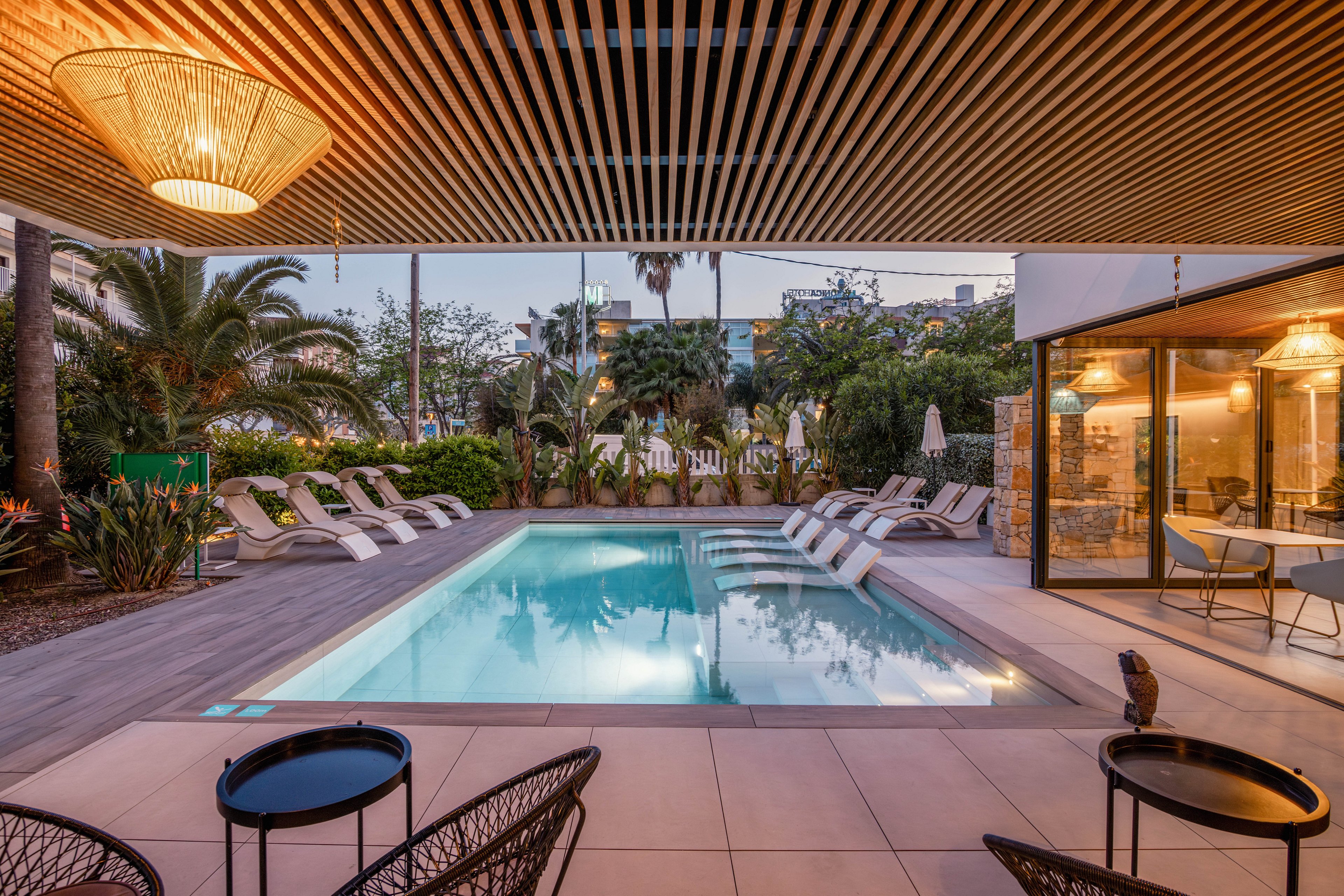 A contemporary outdoor swimming pool is surrounded by cushioned lounge chairs and lush palm trees. The scene is set at dusk, with warm lights glowing from the adjacent glass-walled building. A slatted wooden ceiling and stylish patio furniture create a sophisticated, resort-like atmosphere. The calm water and soft lighting evoke a relaxed, vacation mood.