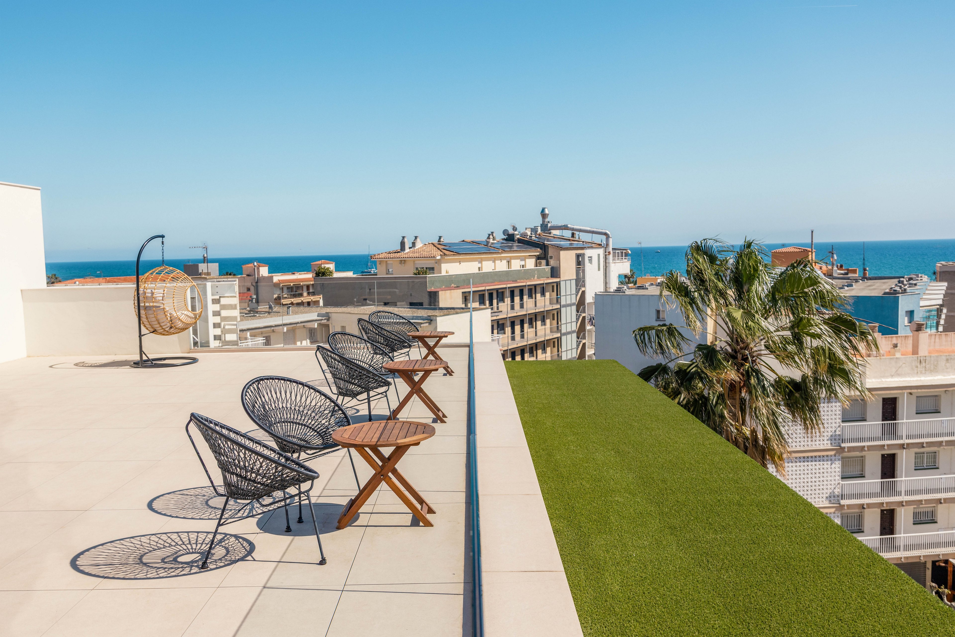 A modern rooftop terrace features several black wire chairs paired with small wooden side tables. The terrace overlooks a coastal cityscape with mid-rise buildings and a clear view of the sea in the distance. A strip of artificial green grass and a tall palm tree add a touch of nature to the urban setting. The bright blue sky and strong sunlight create a clean, minimalist and summery atmosphere.