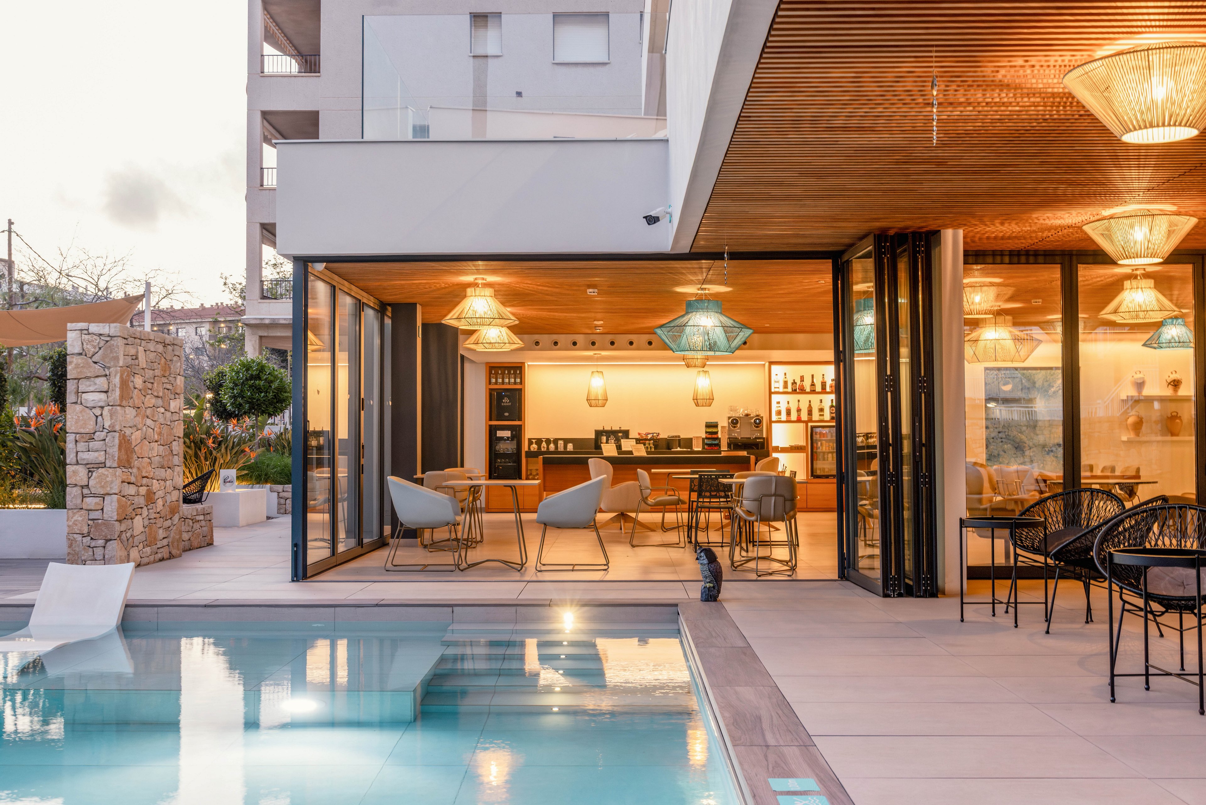 A contemporary poolside lounge area is shown at dusk, featuring a sleek swimming pool in the foreground. Large glass doors open into a warmly lit interior with a bar, dining tables, and comfortable lounge chairs. Pendant lights with decorative shades hang from a wooden ceiling, creating a cozy and inviting atmosphere. The surrounding architecture combines stone, glass, and clean lines for a modern resort-like setting.