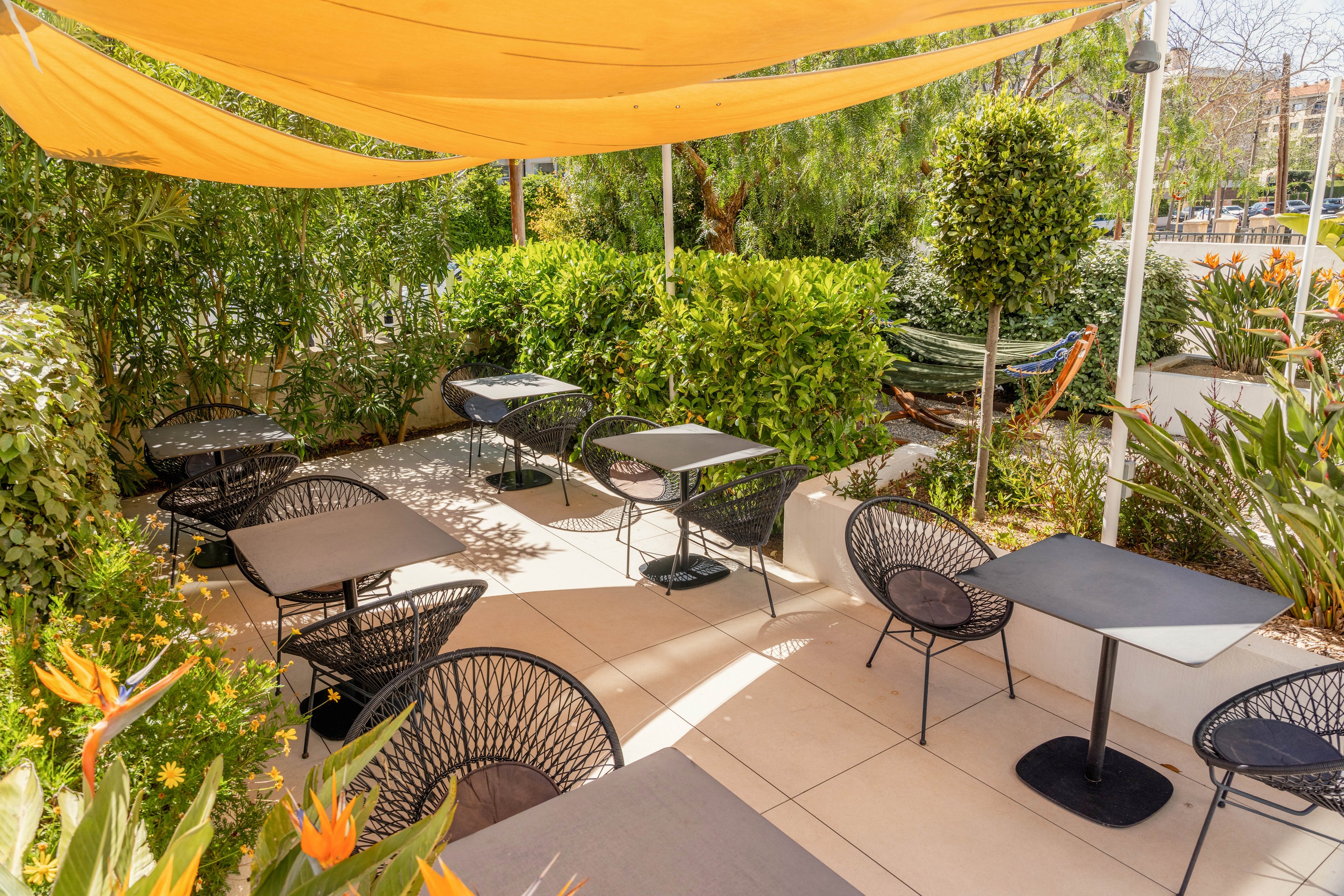 An outdoor café terrace features several empty tables and modern black chairs arranged on a tiled floor. A large yellow fabric canopy provides shade over the seating area. Lush green plants and hedges surround the terrace, creating a cozy garden atmosphere. Bright sunlight and tropical flowers add a warm, inviting feel to the scene.