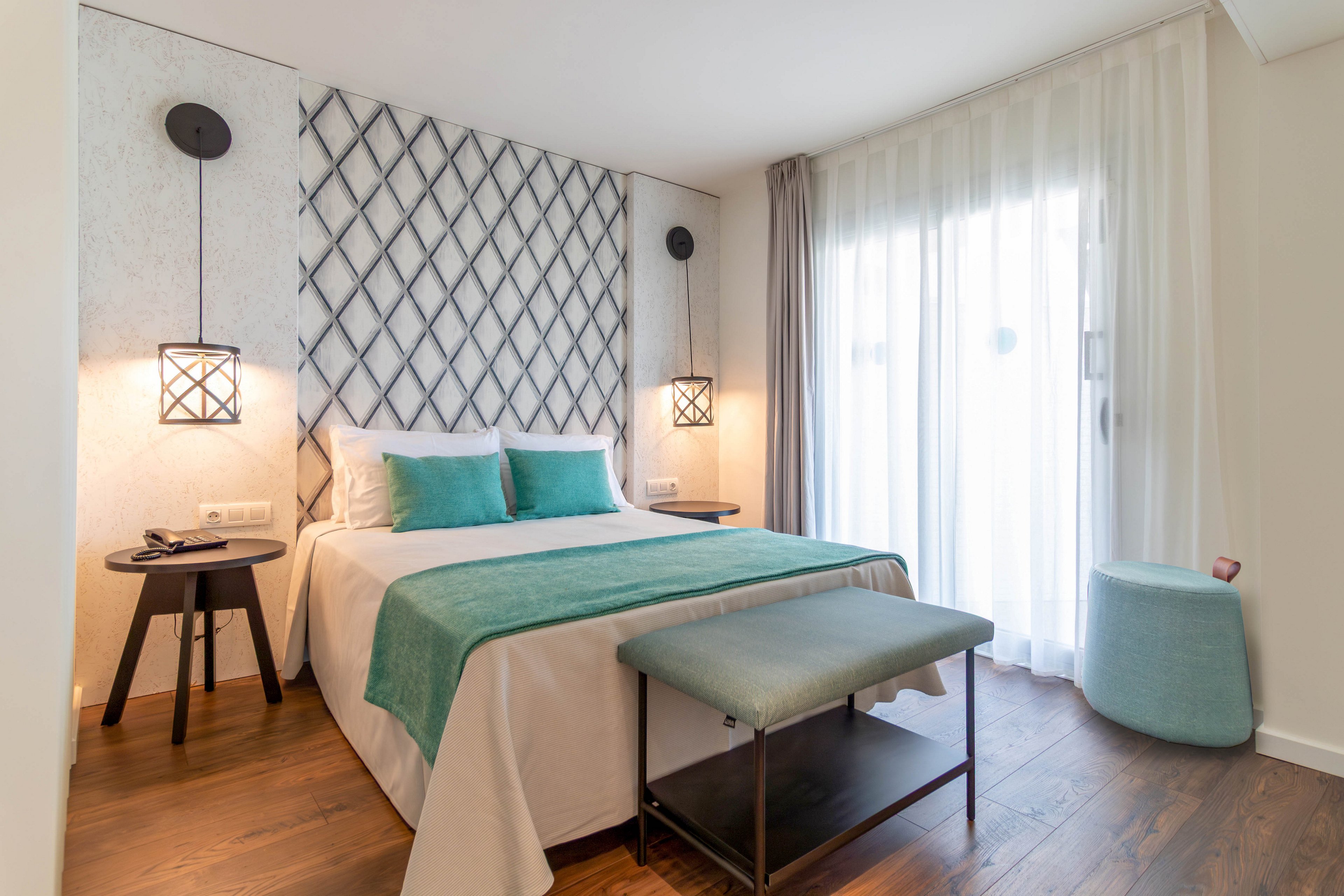 A neatly made double bed with teal pillows and a matching throw is the focal point of this modern hotel room. The space features a decorative lattice headboard, two black bedside tables, and hanging geometric wall lamps. Sheer white curtains cover a large window, allowing soft natural light to fill the room. A teal upholstered bench and round ottoman complement the clean, contemporary decor and warm wooden floor.