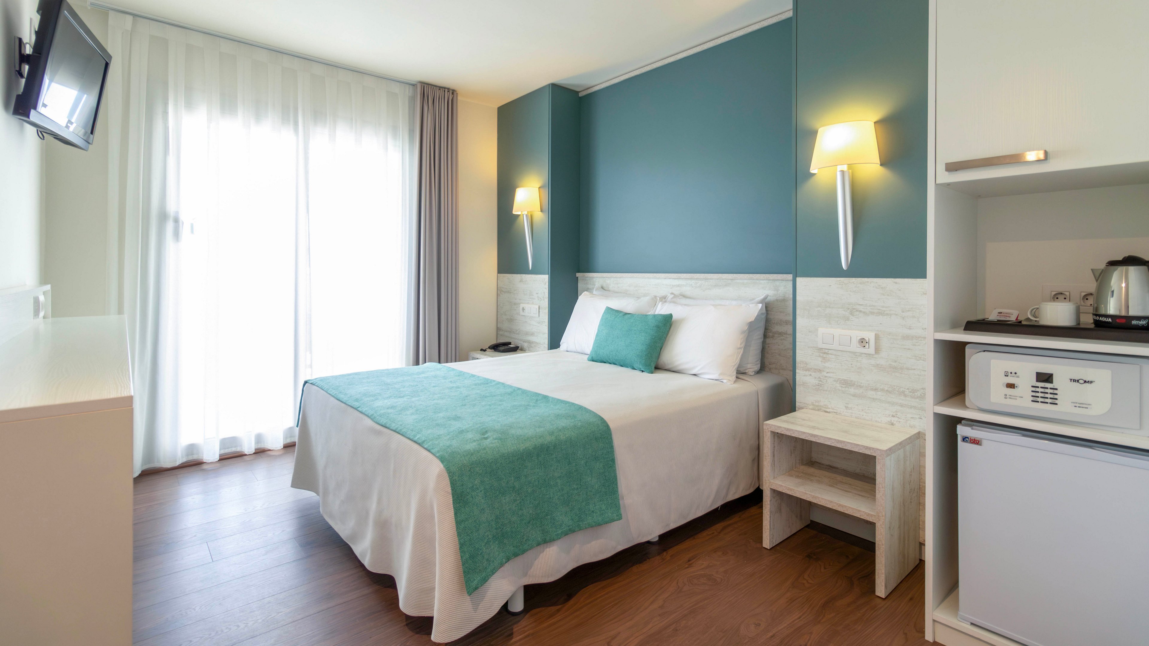 A modern hotel room features a neatly made double bed with white linens and a teal throw and cushion. The space is bright, with natural light streaming through sheer curtains covering a large window. Wall-mounted lamps and a small bedside table frame the bed against a teal accent wall. A compact kitchenette area with a mini fridge, kettle, and cups adds convenience for guests.