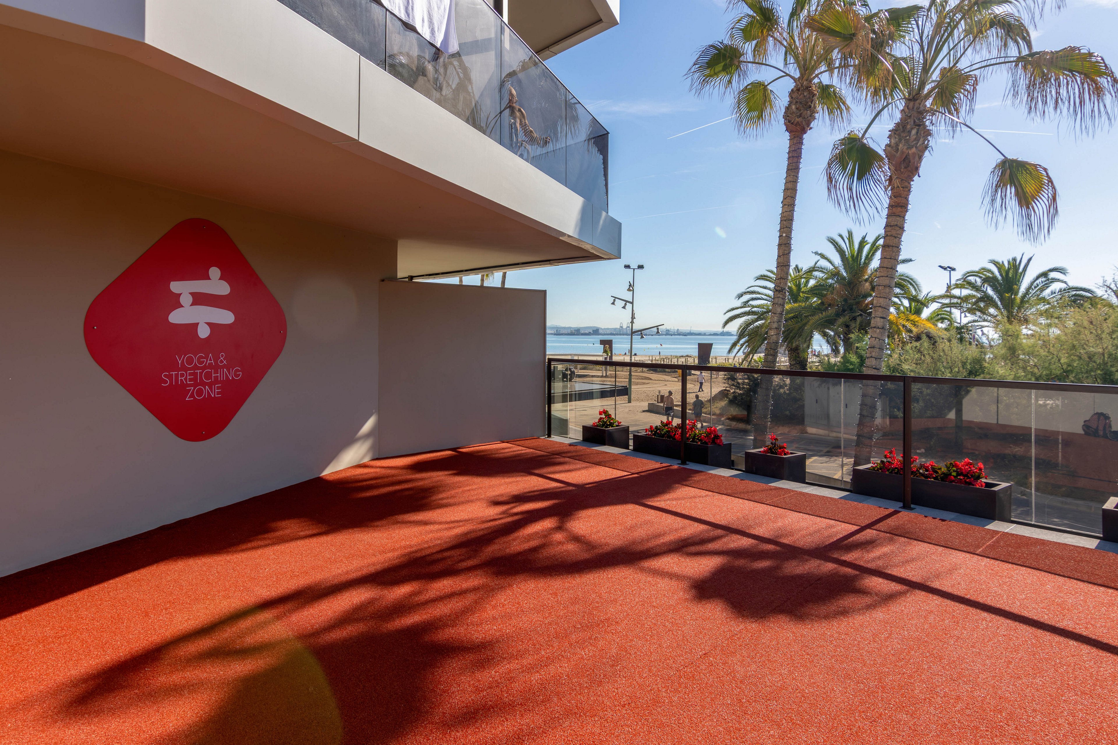 An outdoor yoga and stretching zone is shown on a sunny day near the seaside. The red rubberized floor area is bordered by planters filled with red flowers and a glass railing. Palm trees cast long shadows across the ground, with the beach and ocean visible in the background. A wall sign marks the yoga and stretching zone, adding a subtle focal point to the modern terrace setting.