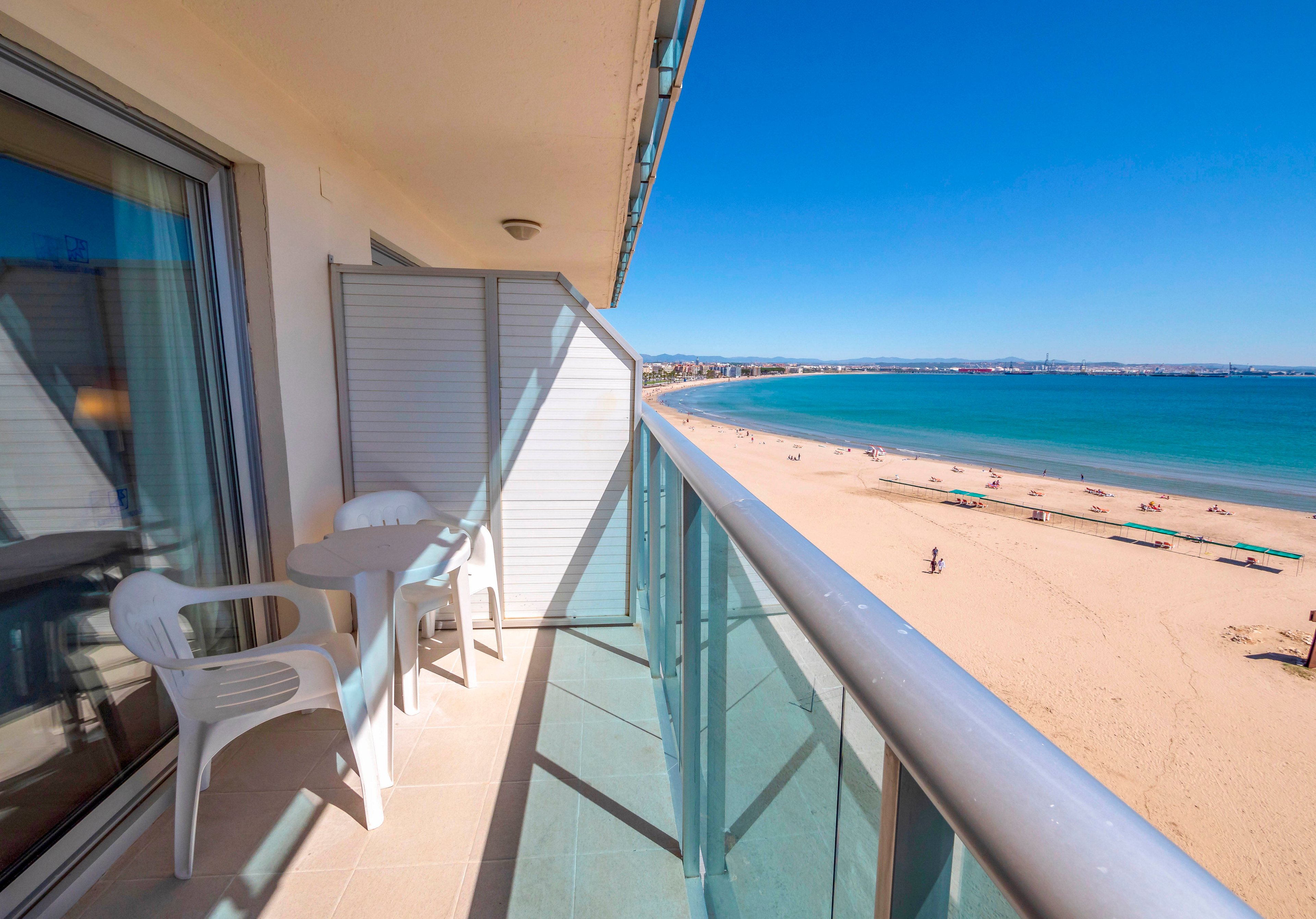 A modern apartment balcony overlooks a wide sandy beach and calm blue ocean. The balcony features a small white table with two plastic chairs beside a glass railing. Bright sunlight and clear blue skies create a warm, inviting atmosphere. People are visible in the distance enjoying the shoreline and water.