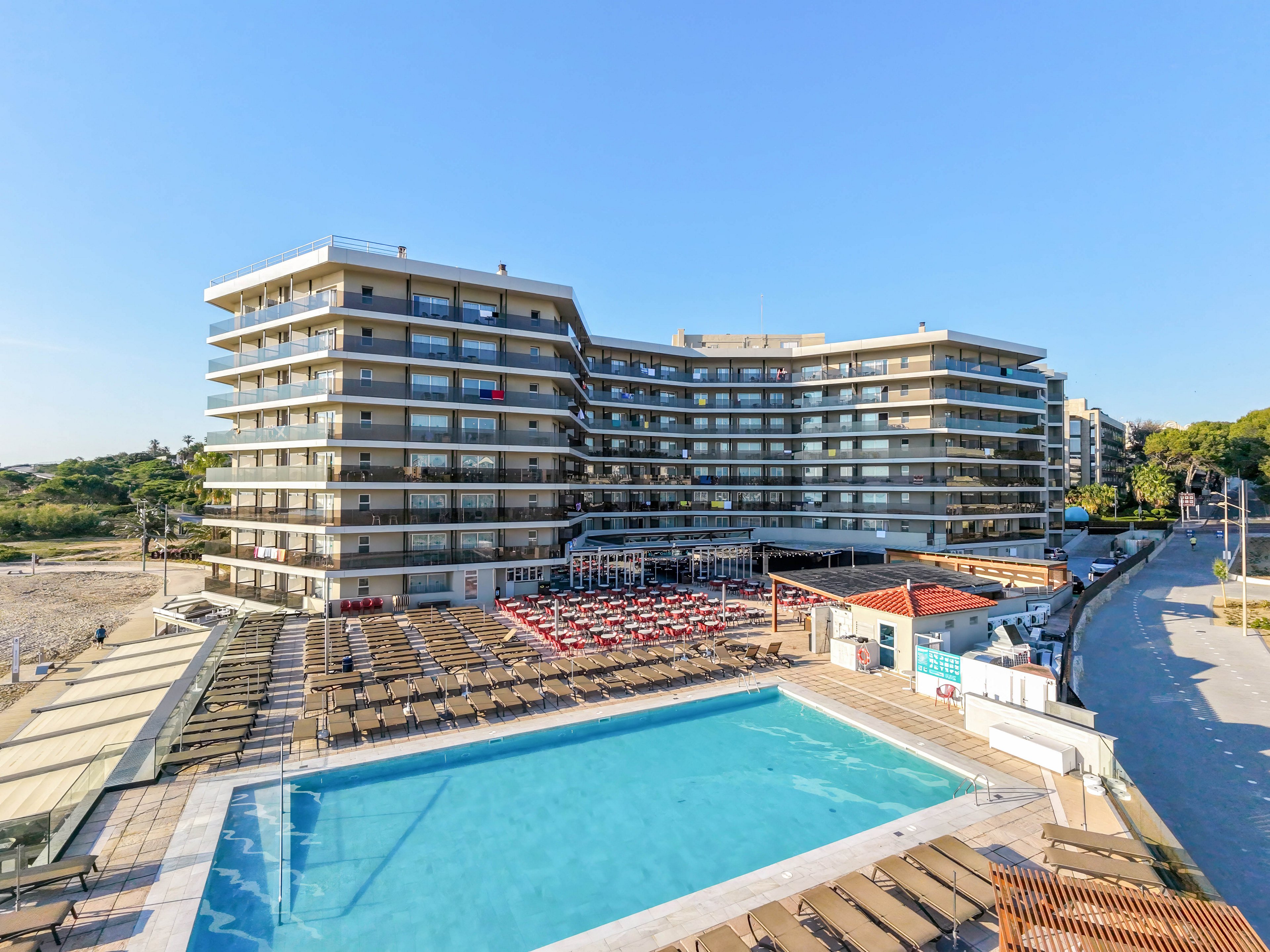 A large multi-story hotel overlooks a spacious outdoor swimming pool. Numerous sun loungers are arranged neatly around the pool, suggesting a resort setting for relaxation. The building faces a sandy beach and open sea under a clear blue sky. Nearby walkways, terraces, and surrounding greenery indicate a coastal vacation destination.