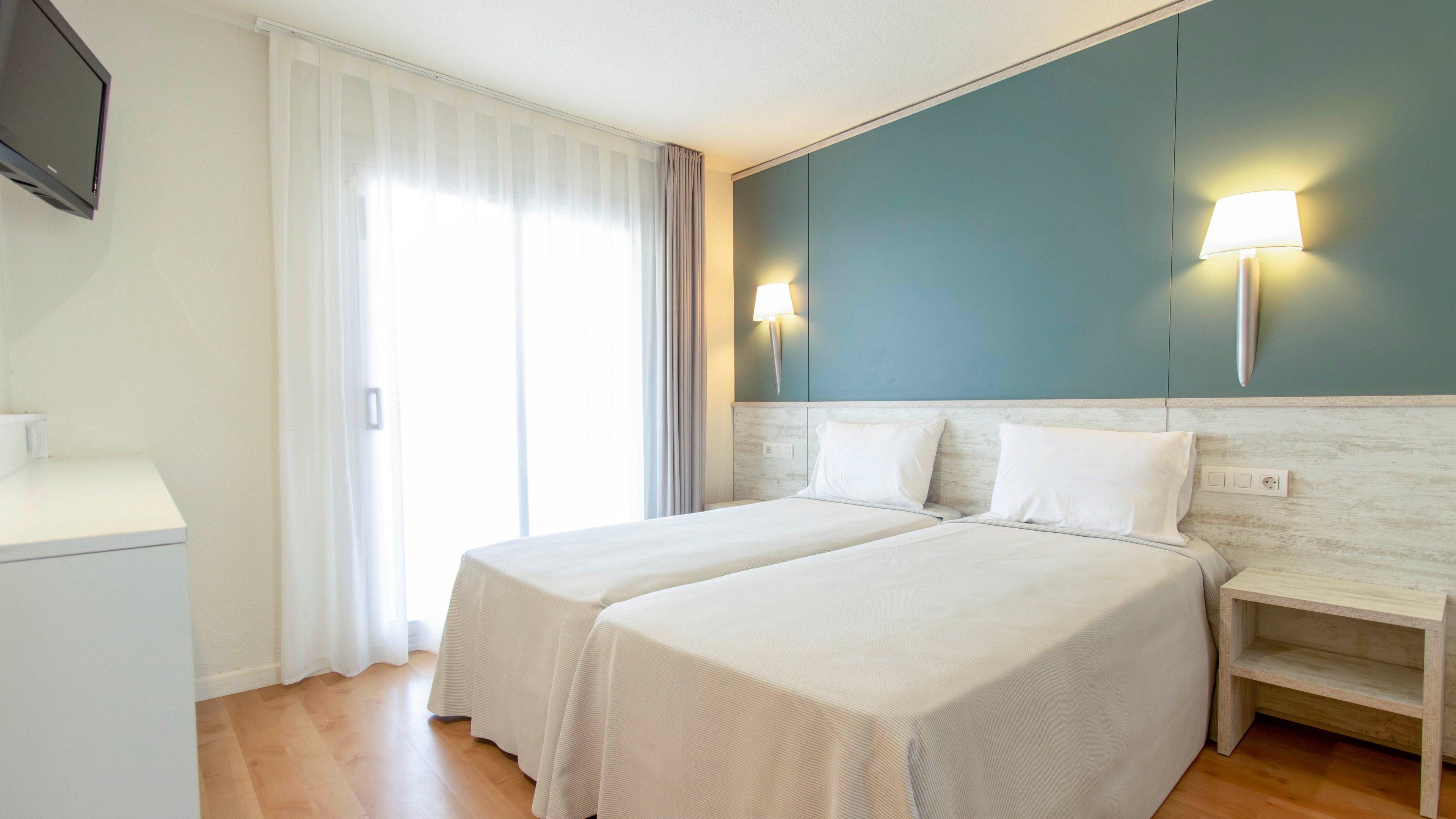 A clean, modern hotel room features two neatly made twin beds with white linens. The space is brightened by natural light coming through large sheer curtains covering a sliding glass door. A teal accent wall behind the beds is complemented by two contemporary wall-mounted lamps. Light wood flooring and minimal furniture, including a bedside table and wall-mounted television, create a simple and inviting atmosphere.