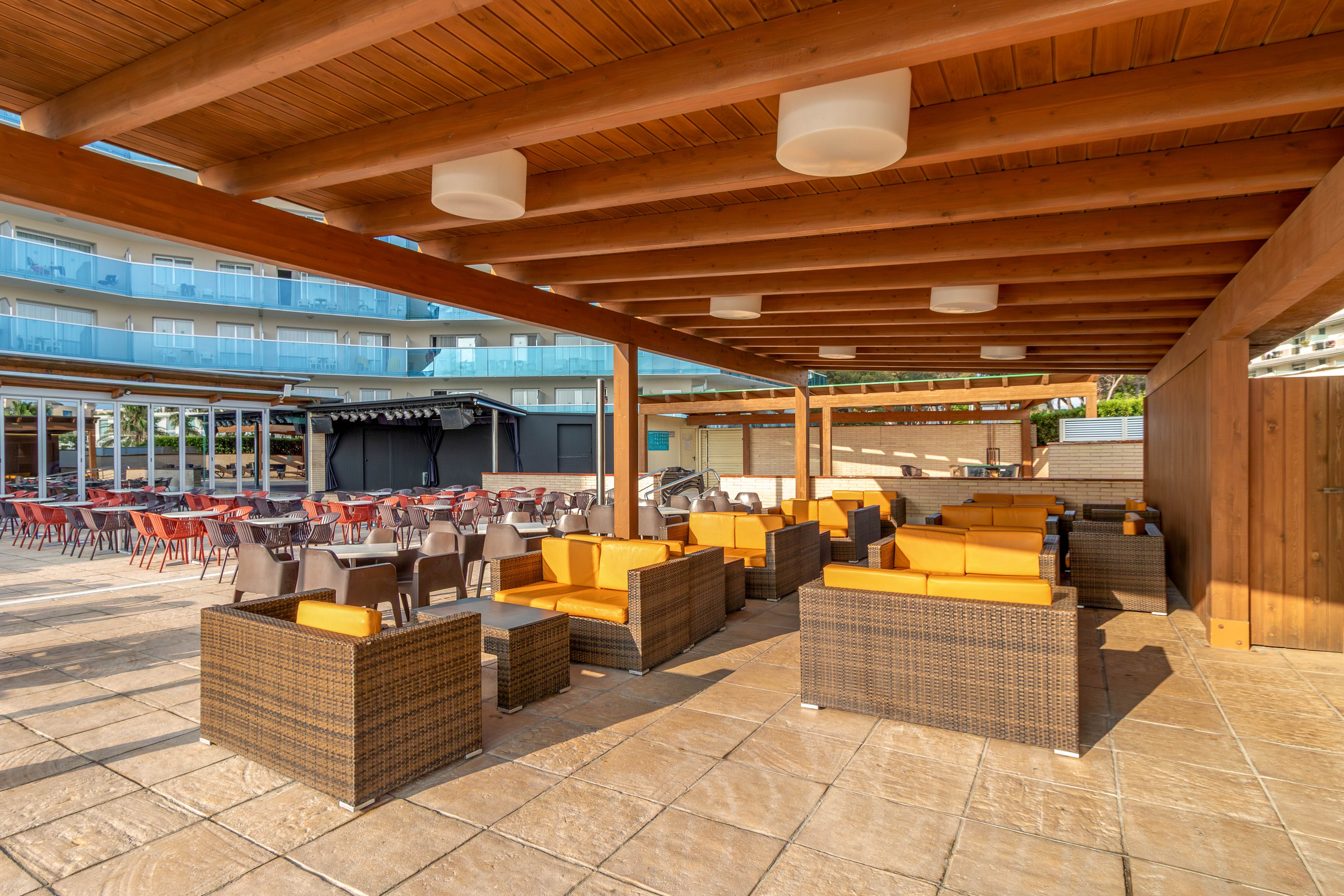An expansive outdoor hotel terrace features wicker lounge furniture with bright yellow cushions under a wooden pergola. In the background, rows of dining tables and chairs are arranged for guests near a bar area. A modern multi-story building with glass balconies overlooks the terrace. The scene is sunlit, creating a warm and inviting atmosphere for relaxation and socializing.