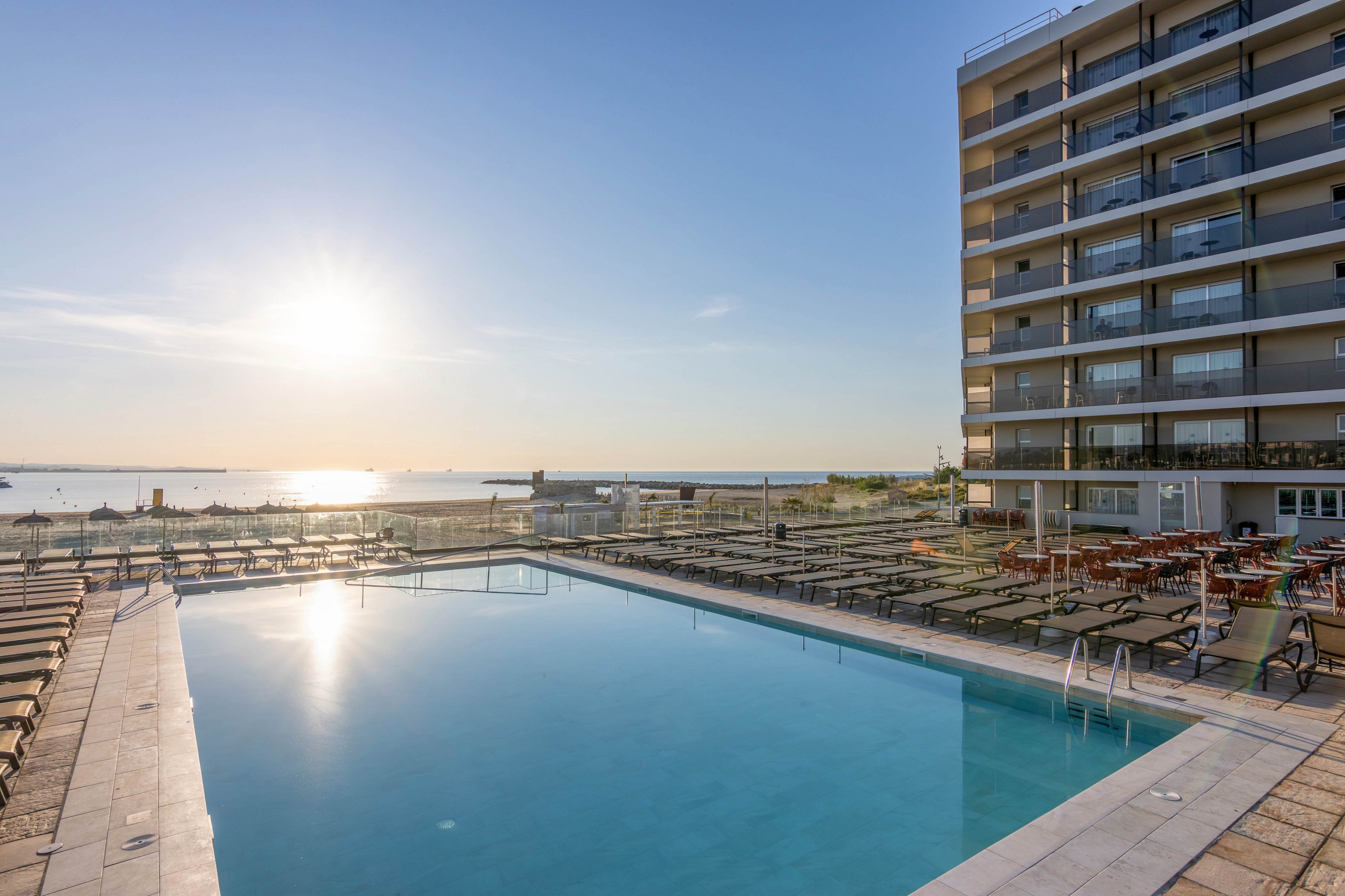 A large outdoor swimming pool reflects the soft light of the rising sun. The poolside area is lined with empty lounge chairs and tables, suggesting an early, quiet hour before guests arrive. A multi-story hotel building stands to the right, overlooking the pool and the distant sea. The horizon and clear sky create a serene, relaxing vacation atmosphere.