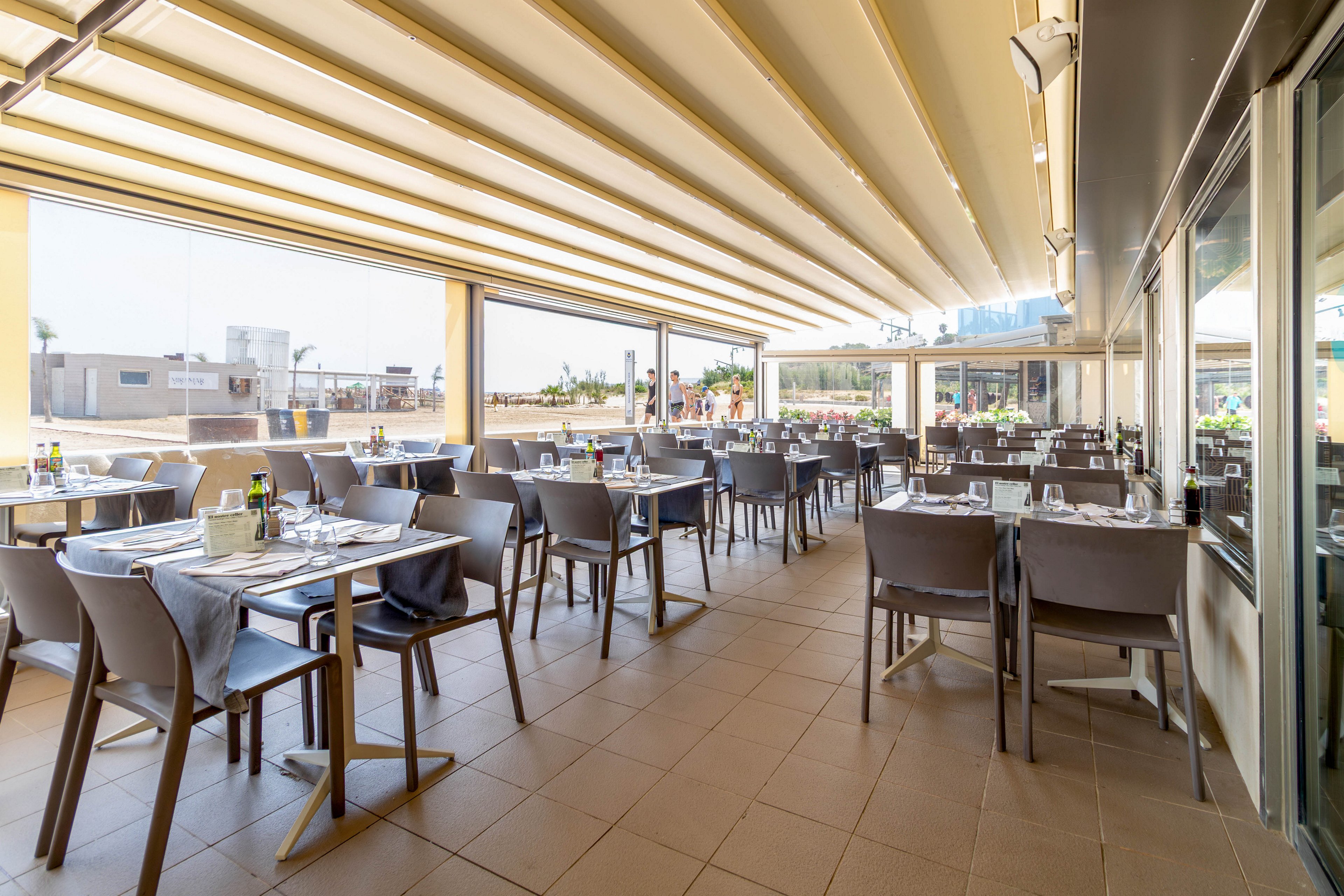 A spacious outdoor restaurant terrace is set with neatly arranged tables and chairs under a modern retractable canopy. Each table is prepared with glassware, cutlery, napkins, and condiments, ready for incoming guests. The bright daylight and open sides reveal a view of surrounding buildings and a relaxed urban environment. The scene feels calm and inviting, suggesting a pleasant dining experience in warm weather.