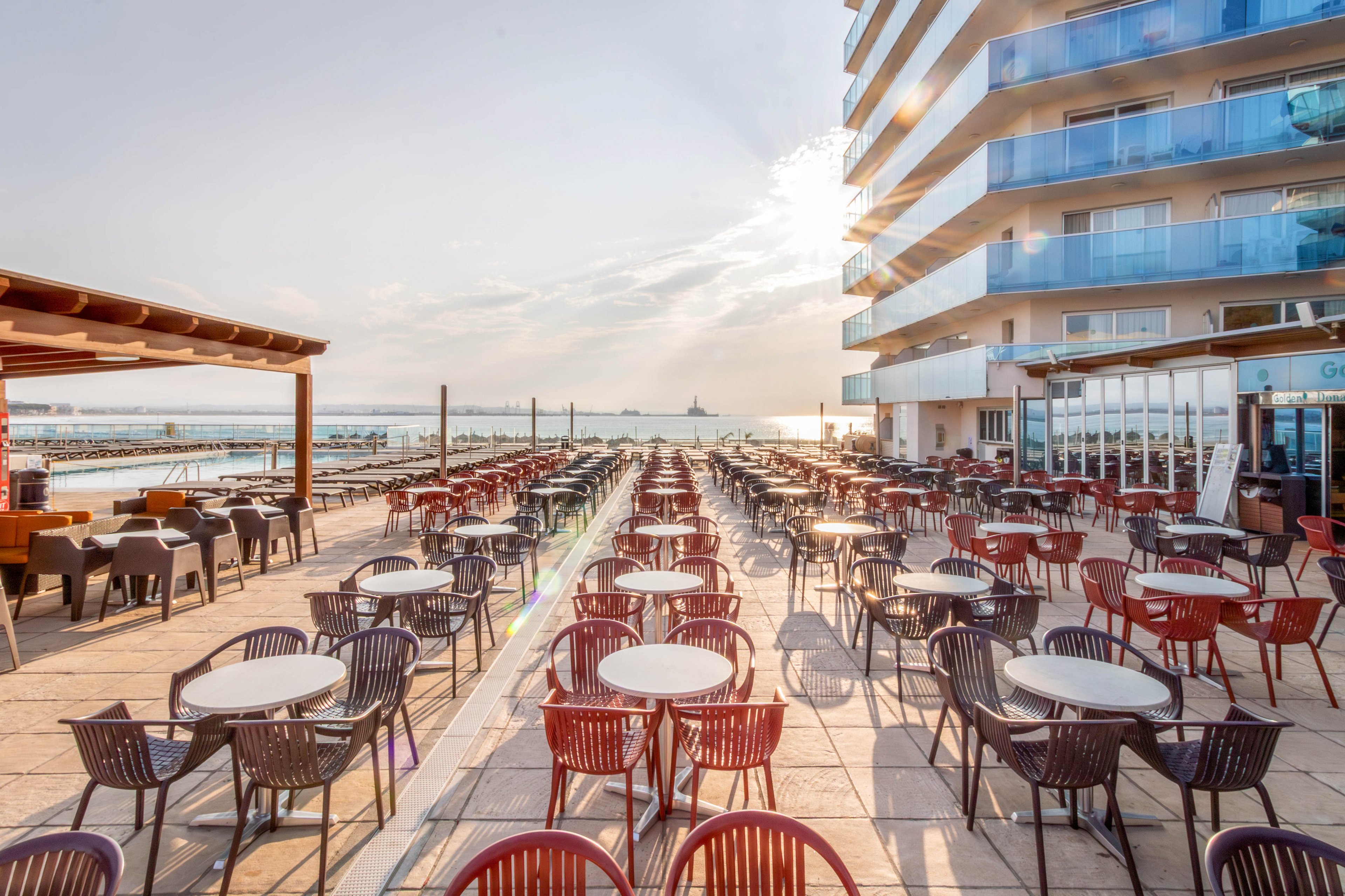 A large outdoor terrace cafe is set with rows of round tables and plastic chairs beside a modern seaside hotel. The scene overlooks a calm body of water with a distant shoreline and structures on the horizon. Morning or late afternoon sunlight creates long shadows and a warm glow across the tiled floor. The area appears ready for guests, emphasizing spacious seating and an open-air coastal atmosphere.