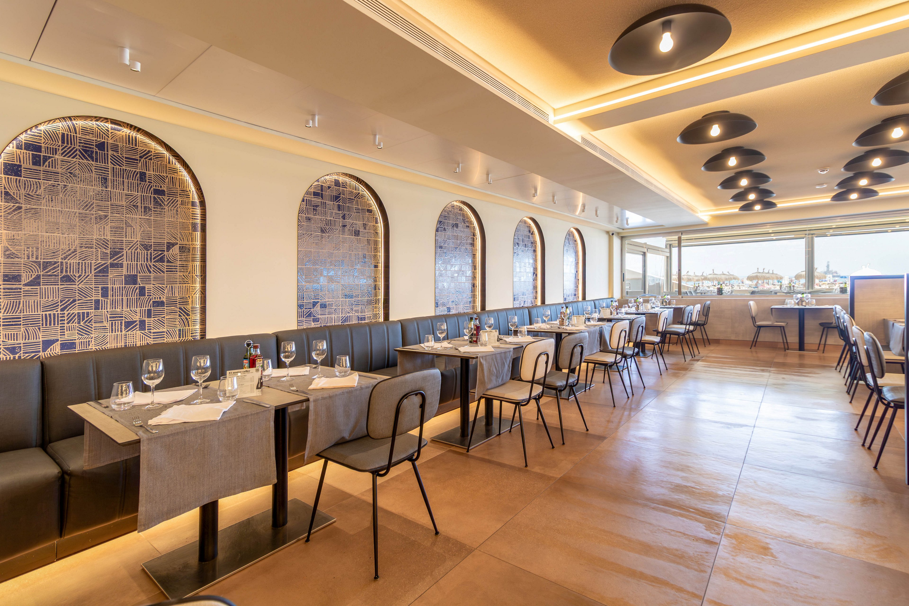A contemporary restaurant interior features neatly arranged tables with place settings and glassware. The space includes cushioned banquette seating along one wall and individual chairs facing concrete-style tables. Arched wall niches with blue geometric tile patterns add decorative interest. Warm ceiling lighting and large windows create an inviting atmosphere with views over the city skyline.