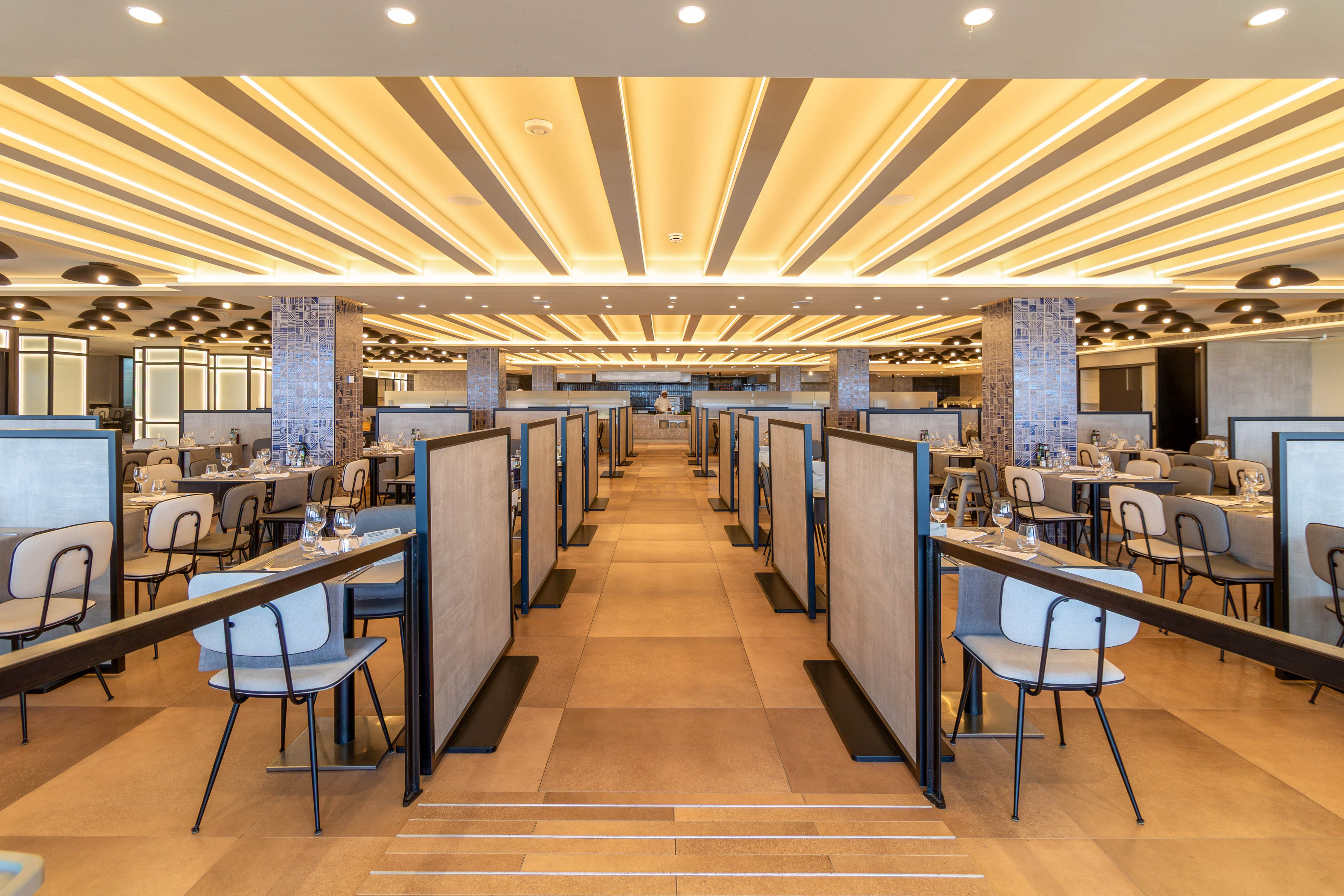A spacious, modern cafeteria interior features long rows of partitioned tables and chairs arranged symmetrically. Warm ceiling lights create a bright, inviting atmosphere across the large open space. The partitions provide semi-private seating areas, and the floor and furniture share a neutral, contemporary palette. No people are present, emphasizing the clean, organized layout of the dining area.