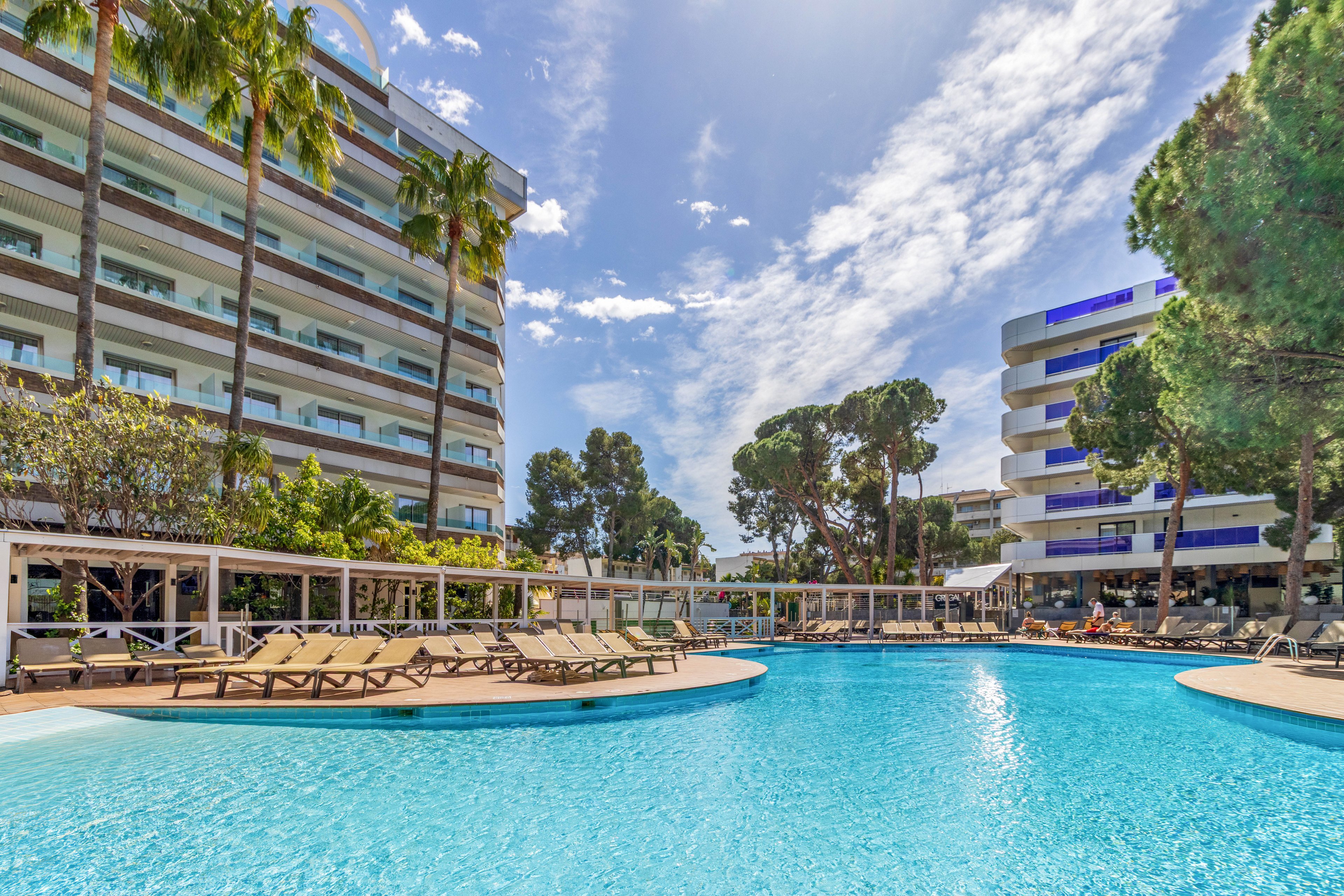 A bright blue swimming pool curves between two modern multi-story hotel buildings. Numerous empty lounge chairs line the poolside under a clear, sunny sky. Tall palm trees and other greenery add a tropical feel to the resort setting. The scene suggests a relaxing vacation atmosphere with warm weather and calm water.