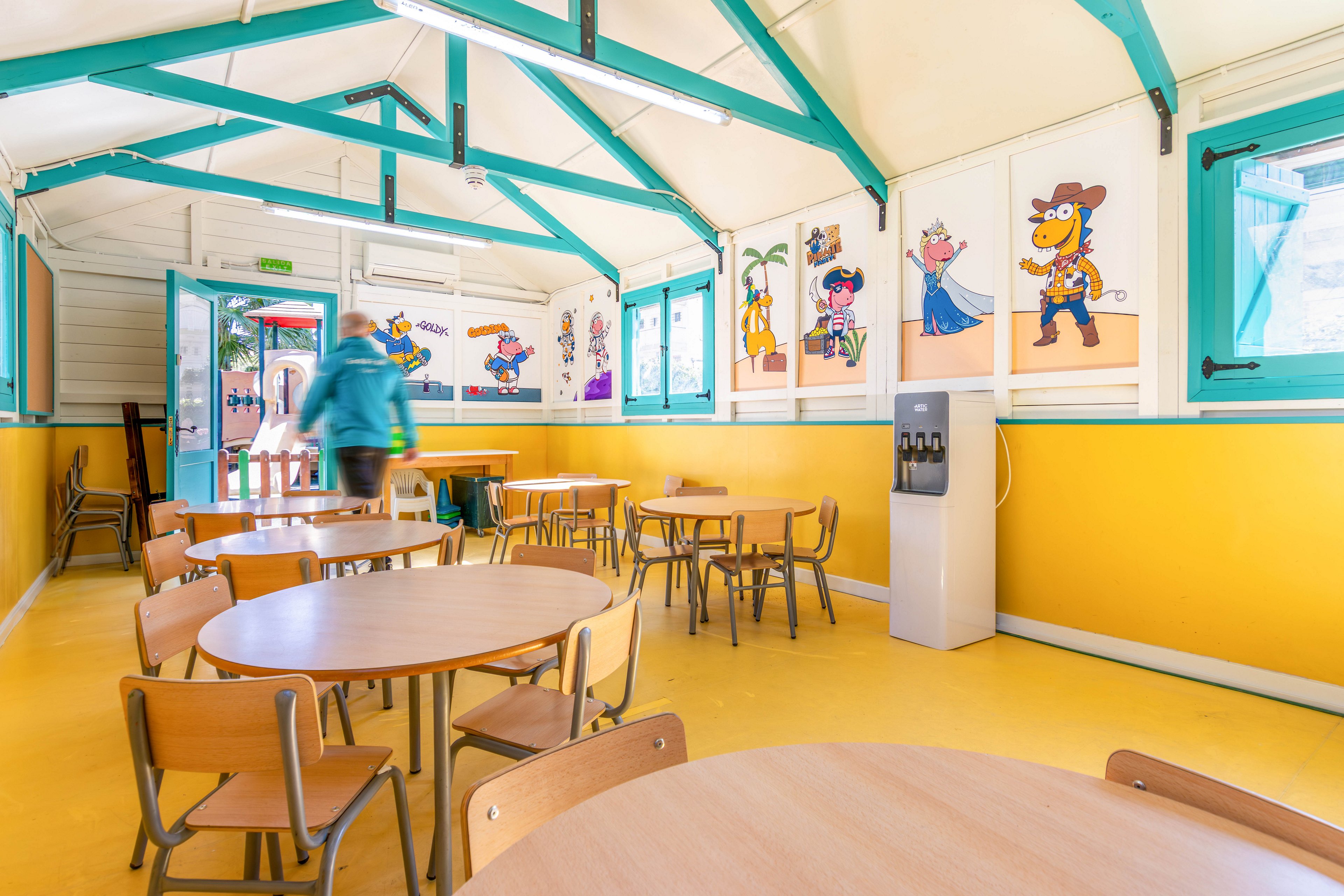 A bright children’s cafeteria features round wooden tables and small chairs arranged neatly in a yellow-floored room. Cartoon murals of playful characters decorate the yellow and white walls, adding a fun and welcoming atmosphere. A person in a teal jacket walks toward the open doorway, bringing a sense of activity to the otherwise empty space. Turquoise beams and window frames contrast with the warm tones, and a water dispenser stands against the wall.