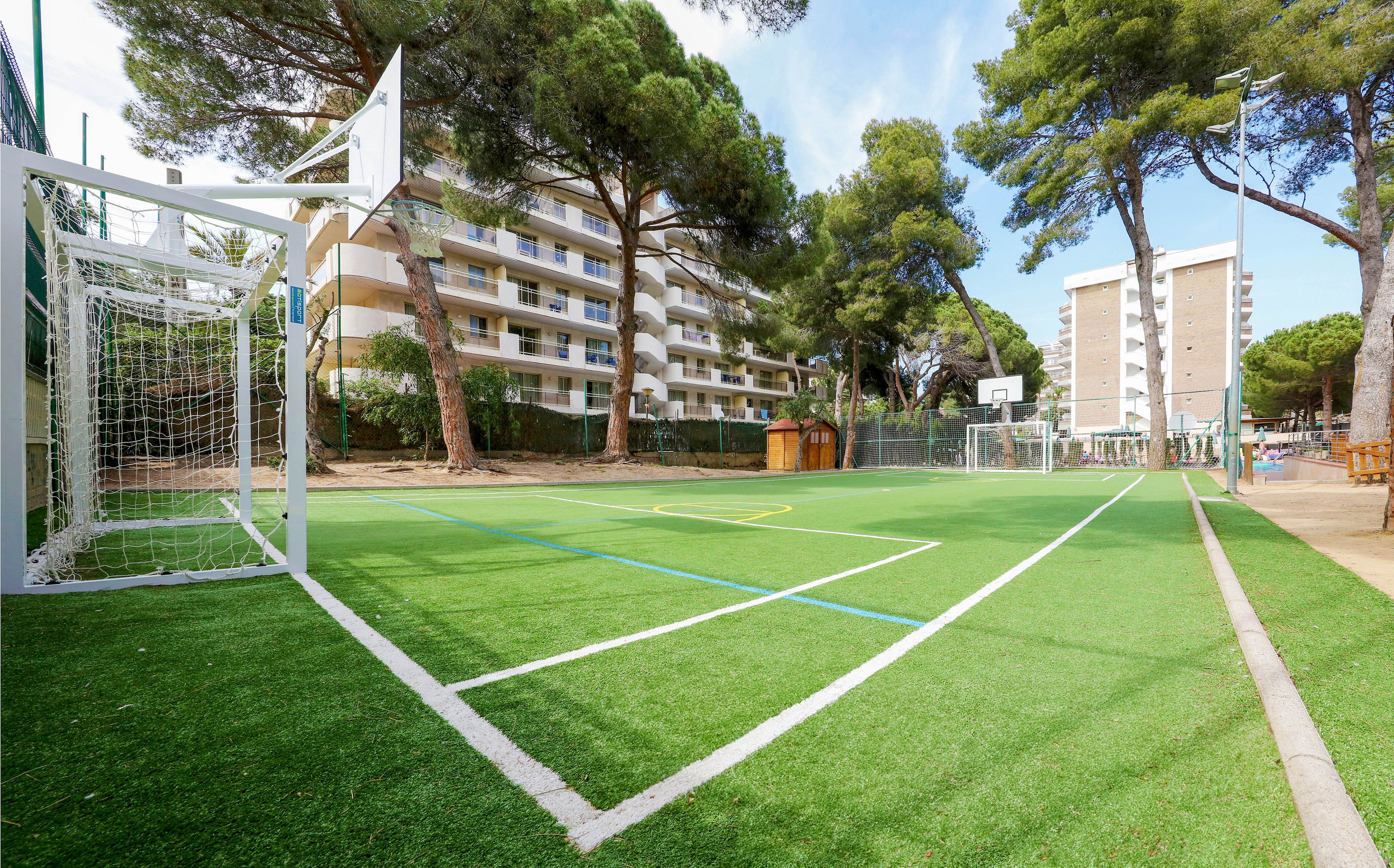 An empty outdoor sports court with artificial turf is shown in a residential area. The court features a soccer goal and a basketball hoop sharing the same space. Tall apartment buildings rise in the background, partially shaded by large pine trees. Bright daylight and clear skies create a clean, inviting atmosphere for recreational activities.