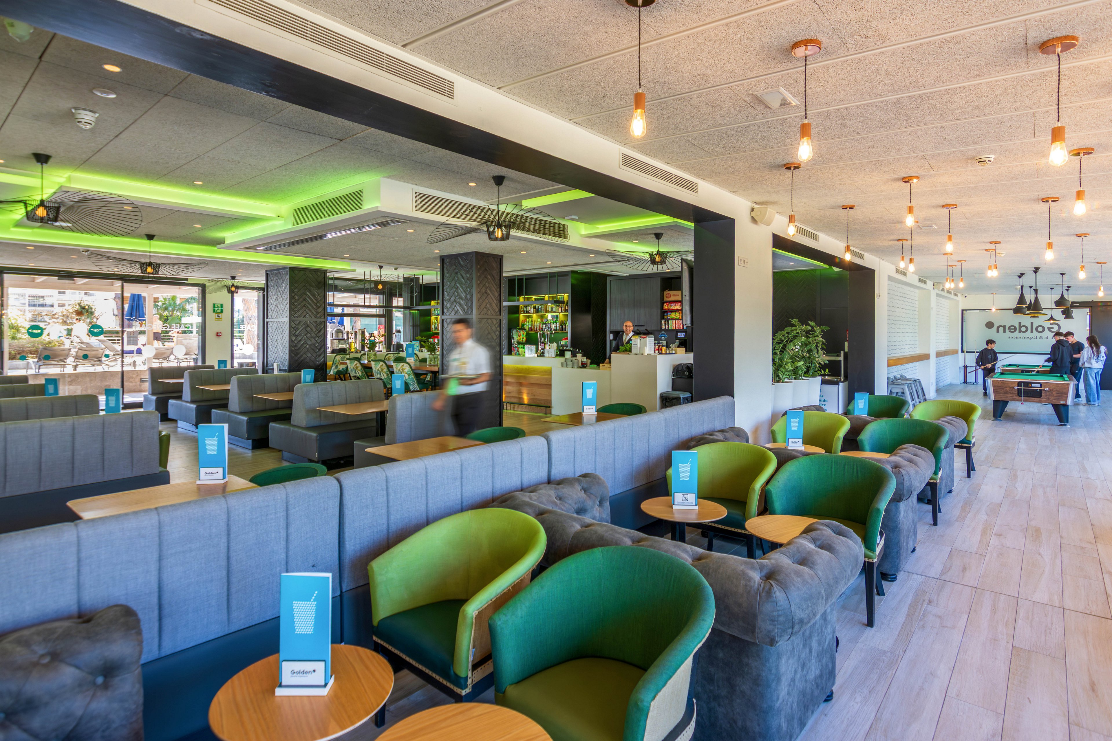A contemporary lounge-style cafe features rows of green and gray armchairs around small wooden tables. The bright interior includes a long bar counter, ceiling pendant lights, and neon green accent lighting. Large windows provide natural light and views of the city outside. A few people are visible in the background, adding a casual, social atmosphere to the space.