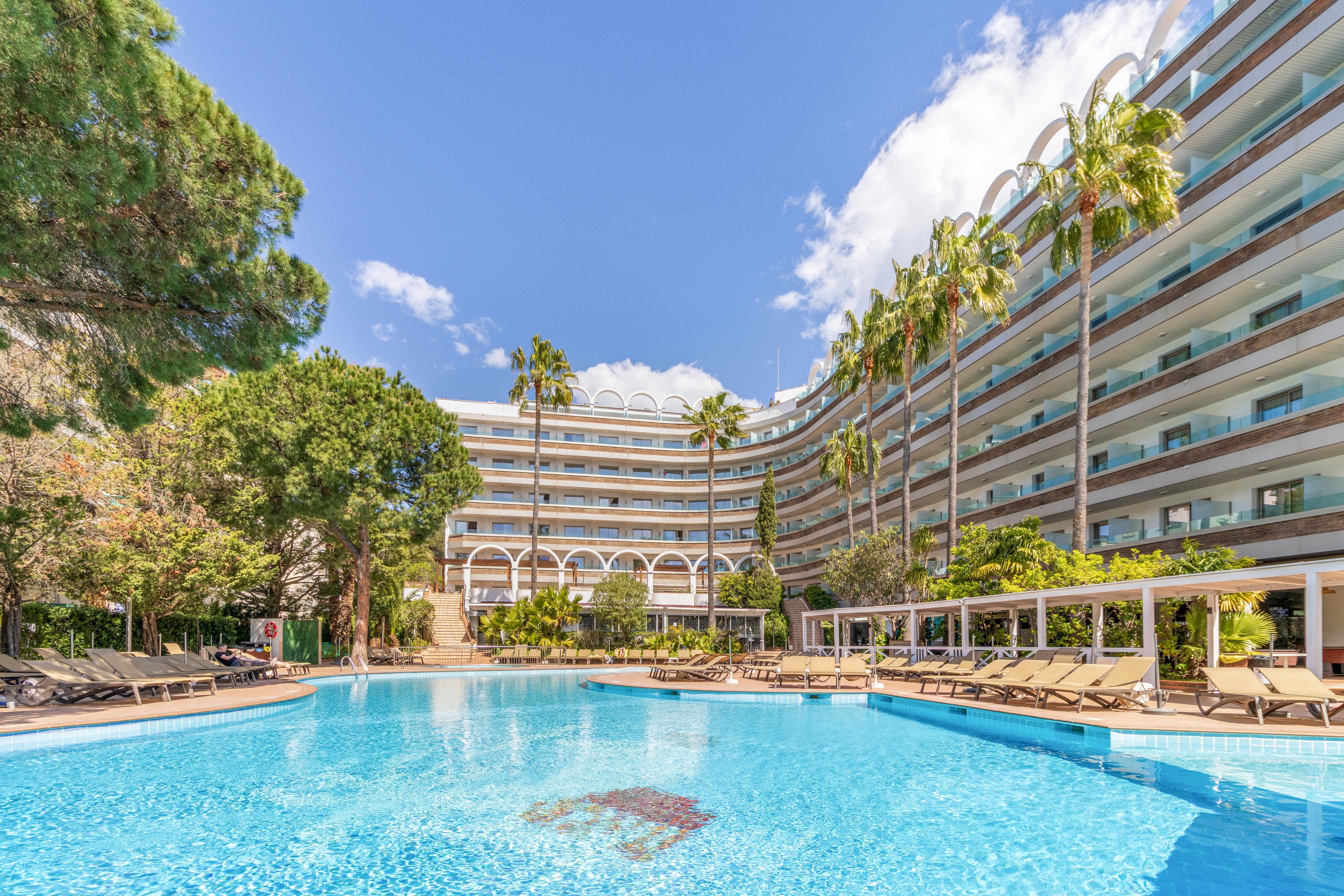 A large outdoor swimming pool sits at the center of a modern resort hotel complex. The multi-story building curves around the pool, with balconies facing the water and tall palm trees adding a tropical feel. Numerous sun loungers line the poolside under clear blue skies with scattered clouds. The bright sunlight and reflections in the water create a relaxed vacation atmosphere.