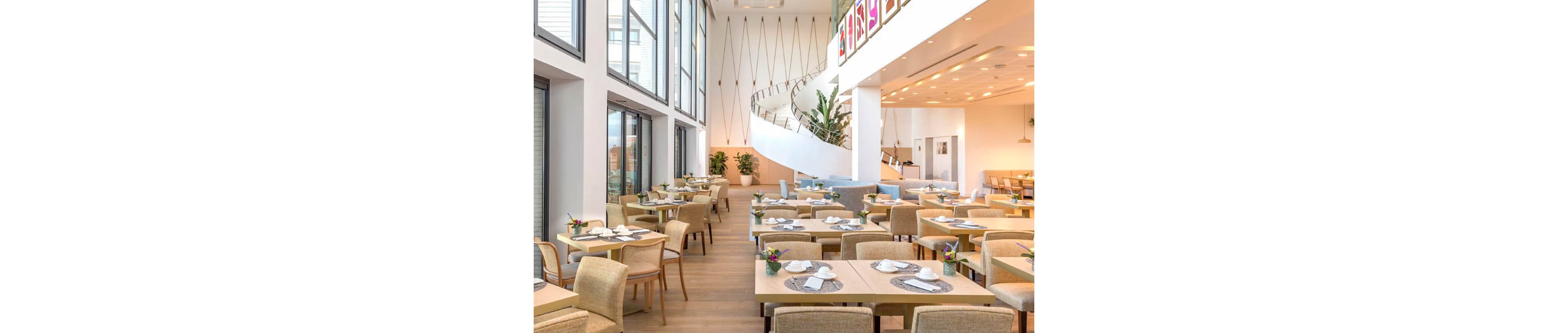 A bright, contemporary dining area is set with neatly arranged tables and chairs. Large floor-to-ceiling windows line one side of the room, filling the space with natural light. A sweeping spiral staircase rises in the background, adding an architectural focal point. Neutral tones, wooden flooring, and small floral centerpieces create a calm, welcoming atmosphere.
