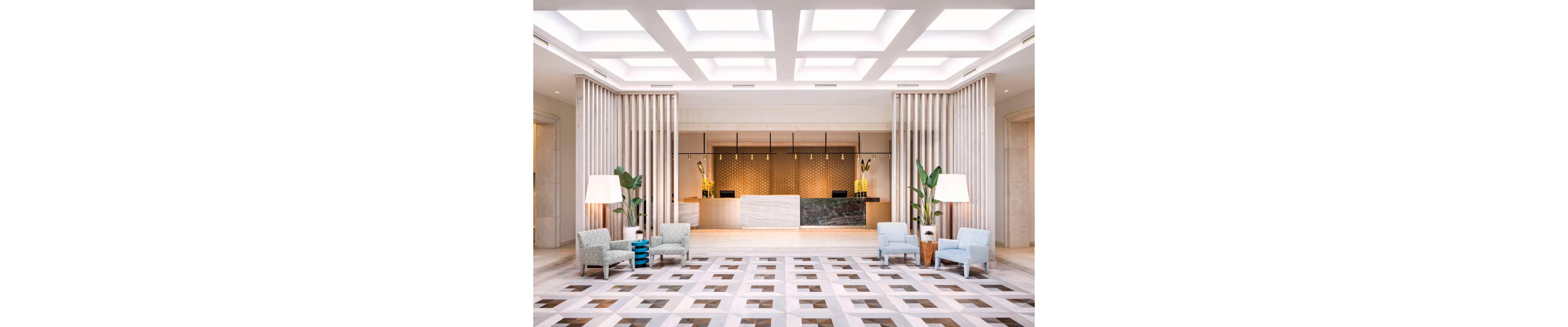A spacious, contemporary hotel lobby features a central reception desk with a textured wall backdrop. The area is framed by vertical wooden slats and large potted plants on both sides. Comfortable armchairs and side tables create seating areas on the patterned geometric floor. Bright ceiling light panels evenly illuminate the clean, minimalist interior design.
