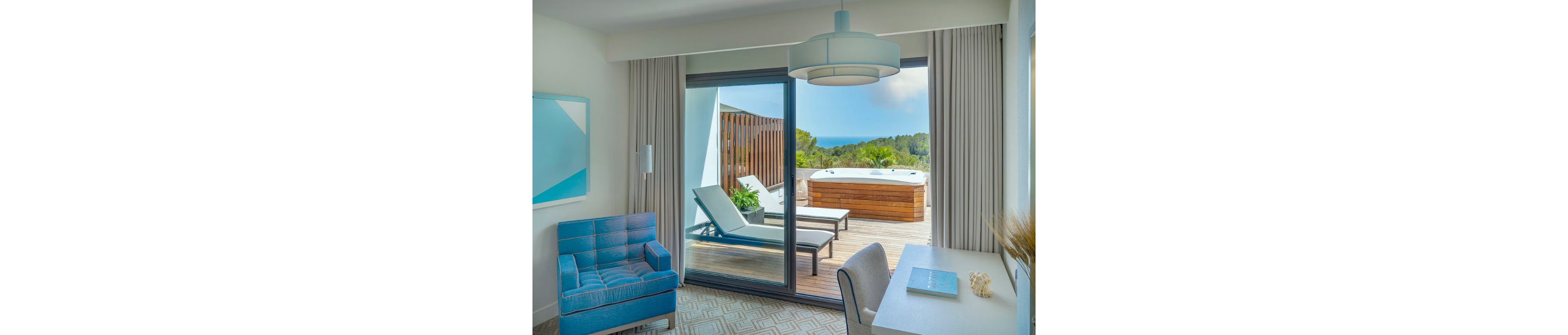 A contemporary hotel room features a blue armchair, white desk, and large sliding glass doors. The doors open to a private terrace with two sun loungers and a wooden hot tub. Beyond the deck, lush greenery and a distant ocean view create a relaxing vacation atmosphere. Soft neutral tones with blue accents give the space a clean, modern feel.