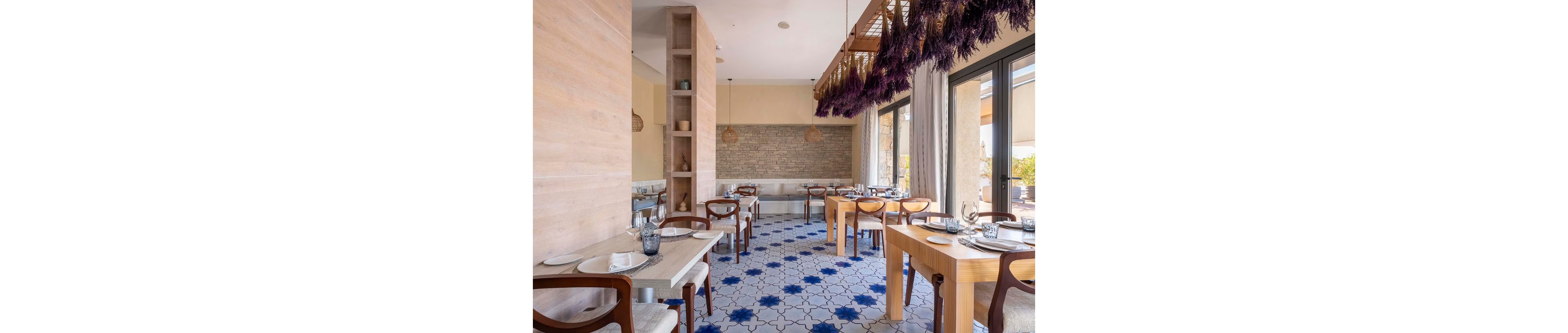 Contemporary restaurant dining room with wooden tables and chairs is shown in natural daylight. Large windows on the right side let in sunlight and views of the outdoors. The floor features a geometric tile pattern in white and blue tones, adding visual interest. Decorative hanging elements and neutral walls create a relaxed, stylish atmosphere with neatly set tables and glassware.