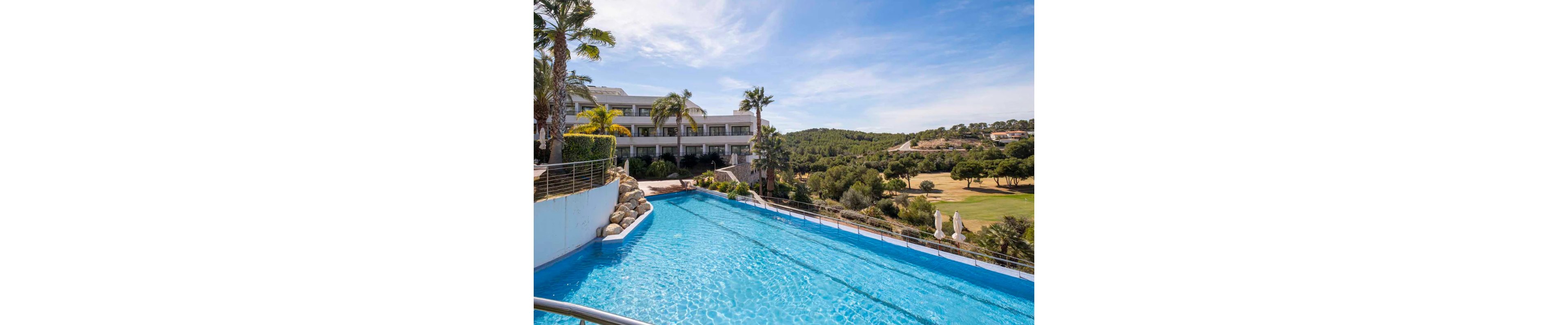 A bright blue infinity pool stretches toward a landscaped golf course and forested hills under a clear sky. A modern multi-story resort building with balconies stands to the left, framed by palm trees and stone landscaping. Sun loungers and umbrellas are visible near the fairway, suggesting a relaxing vacation setting. The overall scene conveys leisure, sunshine, and upscale outdoor amenities.