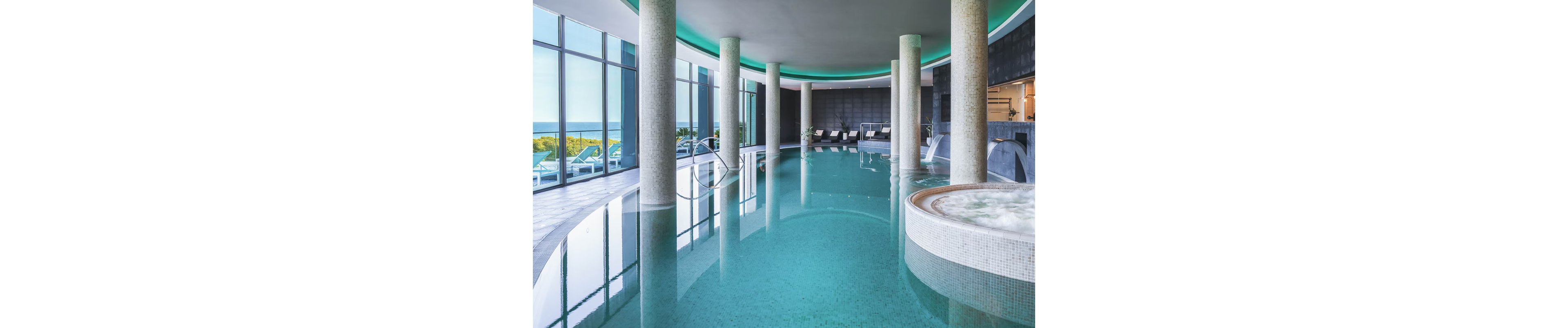 A contemporary indoor spa pool features turquoise water surrounded by tall cylindrical columns and a curved ceiling with accent lighting. Lounge chairs line the far side of the pool near large floor-to-ceiling windows overlooking the ocean. A round whirlpool or hot tub sits in the foreground with mosaic tile detailing. The overall atmosphere is calm and luxurious, suggesting a high-end wellness or resort setting.