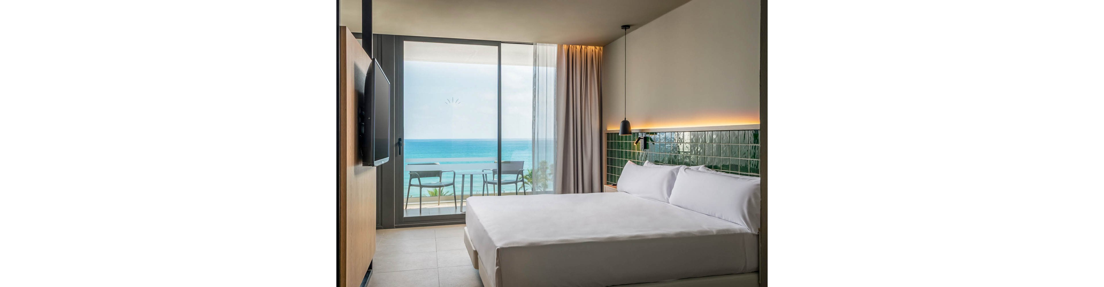 A contemporary hotel room features a neatly made bed and a wall-mounted television. The space opens onto a balcony with two chairs and a table, overlooking a serene blue ocean. Natural light fills the room through large glass doors, highlighting minimalist decor and green tiled accents. The mood is calm and inviting, with neutral tones and soft lighting.