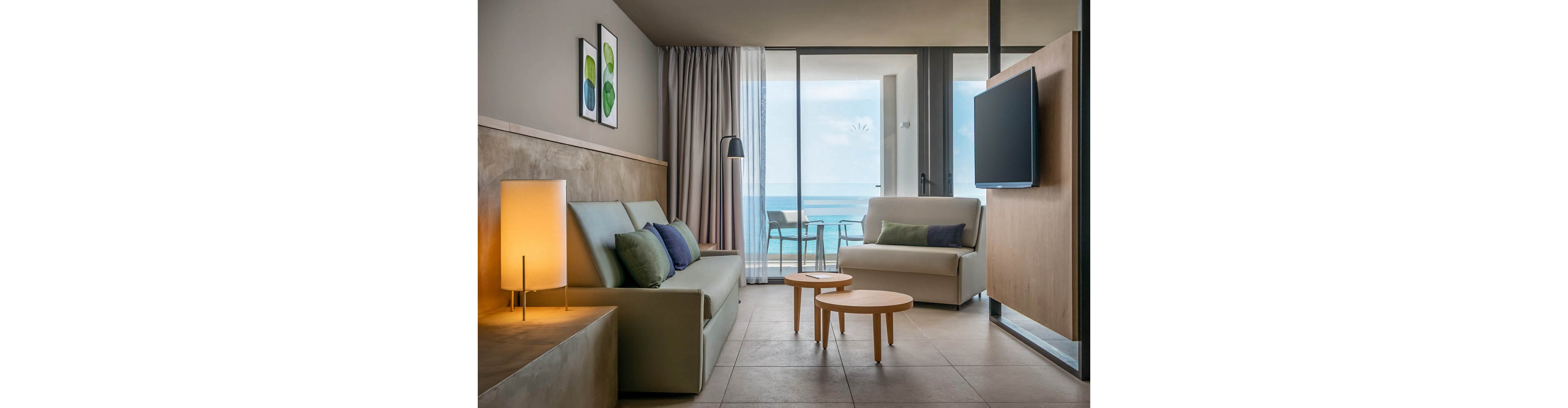 A contemporary hotel suite features a minimalist sofa, armchair, and two round coffee tables. The room is illuminated by soft lighting and decorated with abstract wall art. Large glass doors open to a balcony overlooking the ocean, creating a serene and airy atmosphere. Neutral tones and pops of green and blue enhance the tranquil mood.