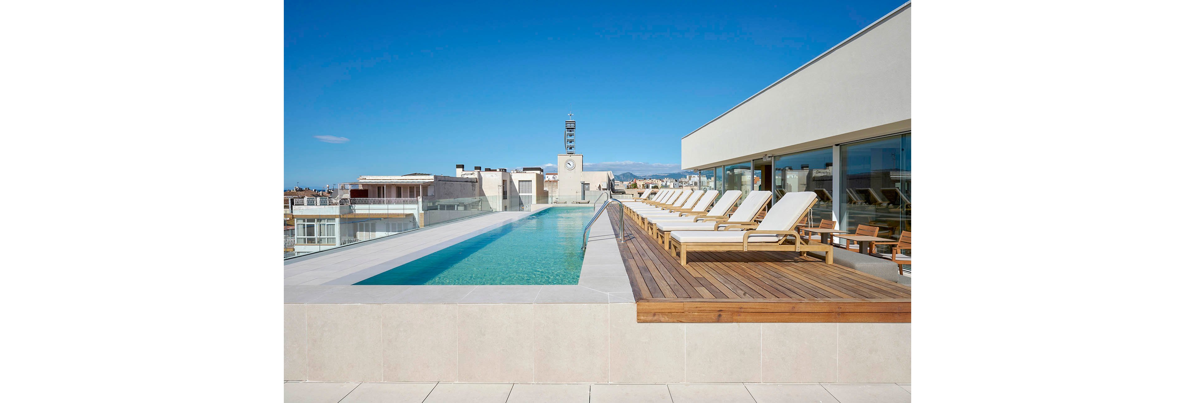 A sleek rooftop infinity pool stretches along the edge of a contemporary building. White cushioned sun loungers line the wooden deck beside the pool, facing the city skyline. Glass walls on the right reflect the bright blue sky and minimalist architecture. The scene suggests a luxurious, tranquil outdoor relaxation area in an urban setting.